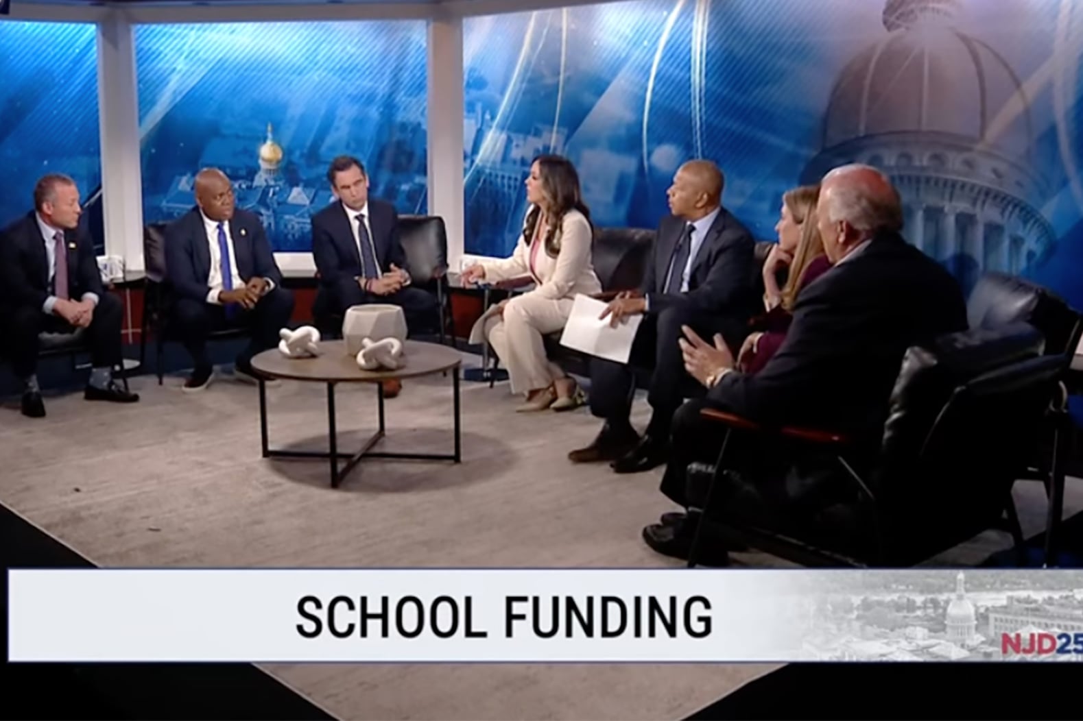 Seven people in business clothing sit at couches on a TV news set.