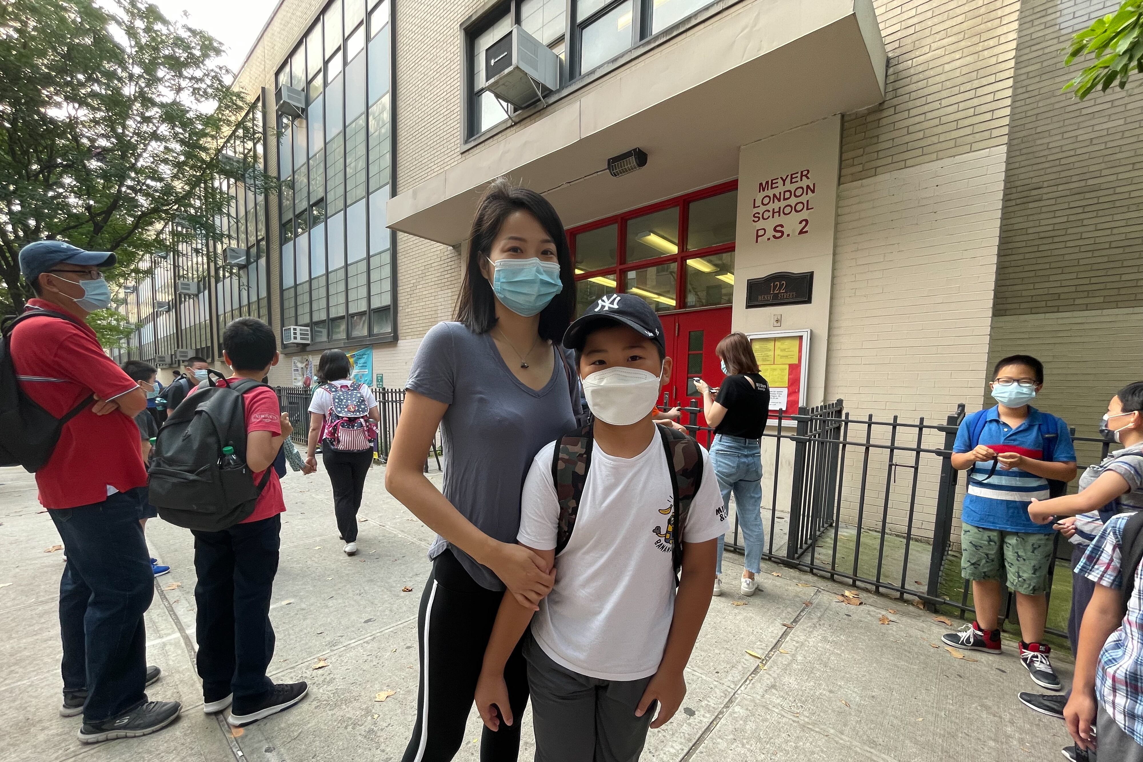A woman and her son stand in front of a school building among other parents and children for a summer program, all wearing protective masks.