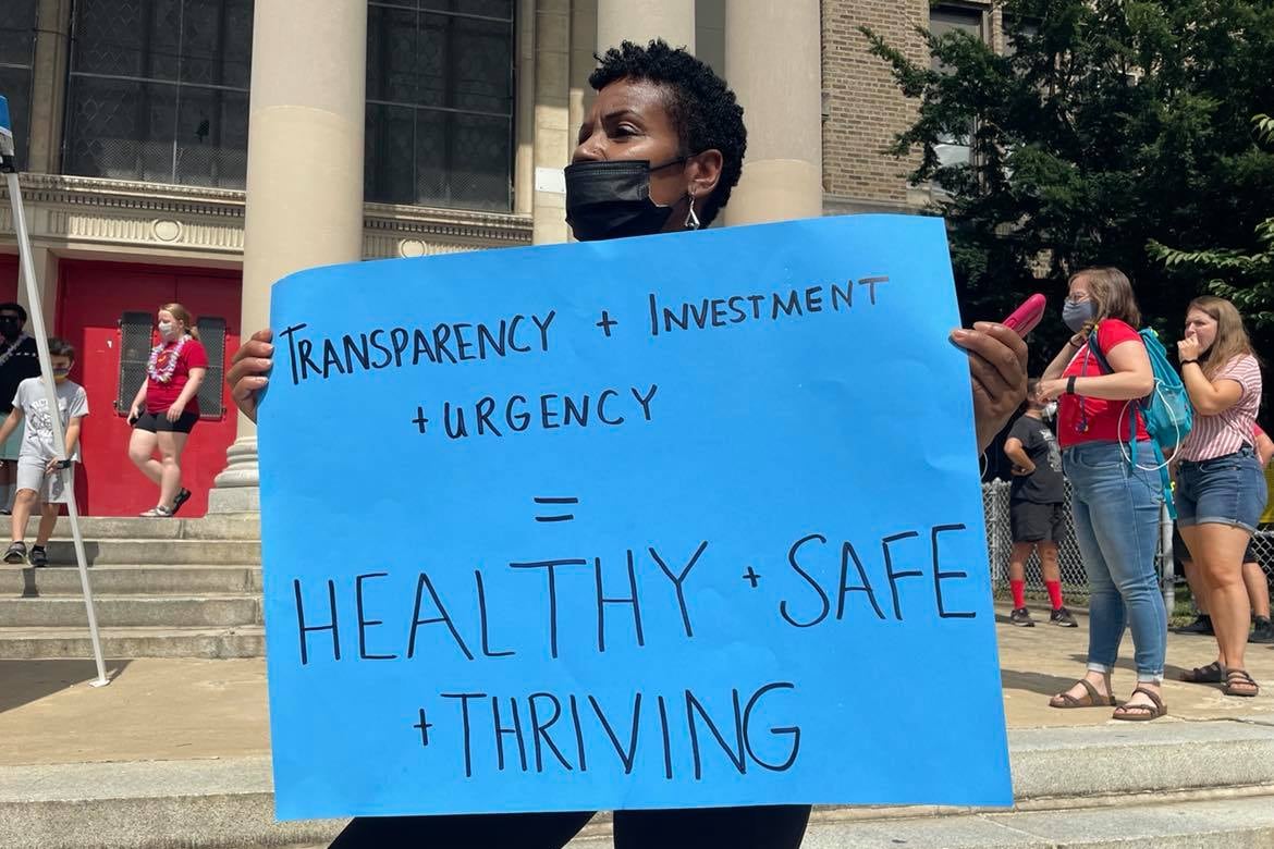A woman holds a large blue sign in front of Science Leadership Academy at Beeber in Wynnefield.