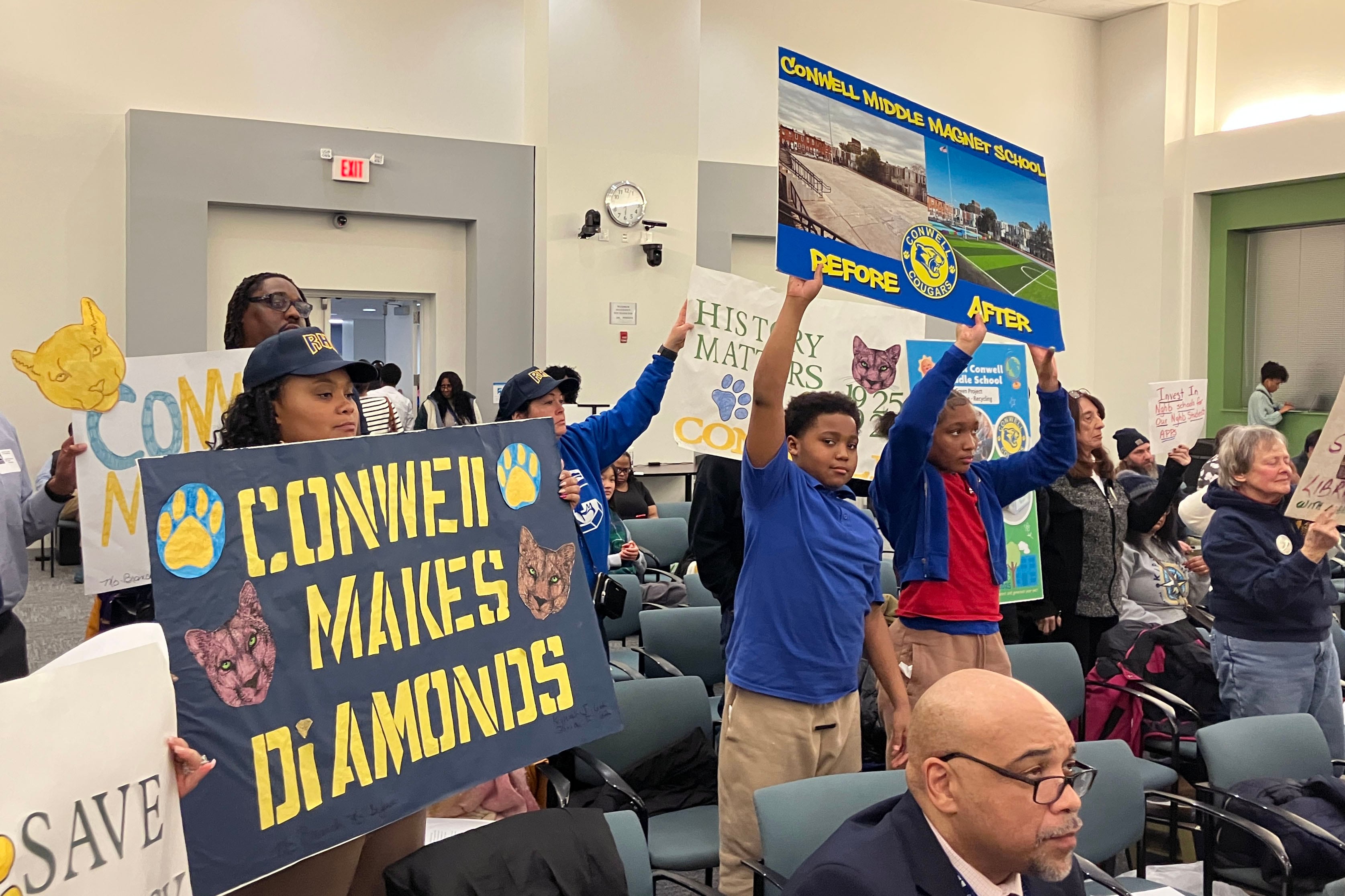 A photograph of children and adults holding protest signs in a conference room.