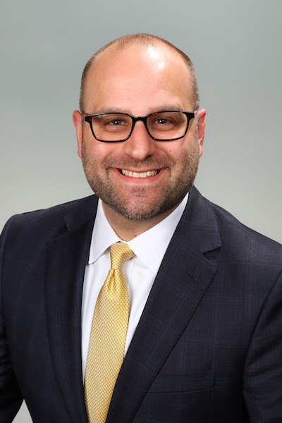 A man with black-framed glasses wearing a dark suit with a yellow tie smiles for a professional headshot.