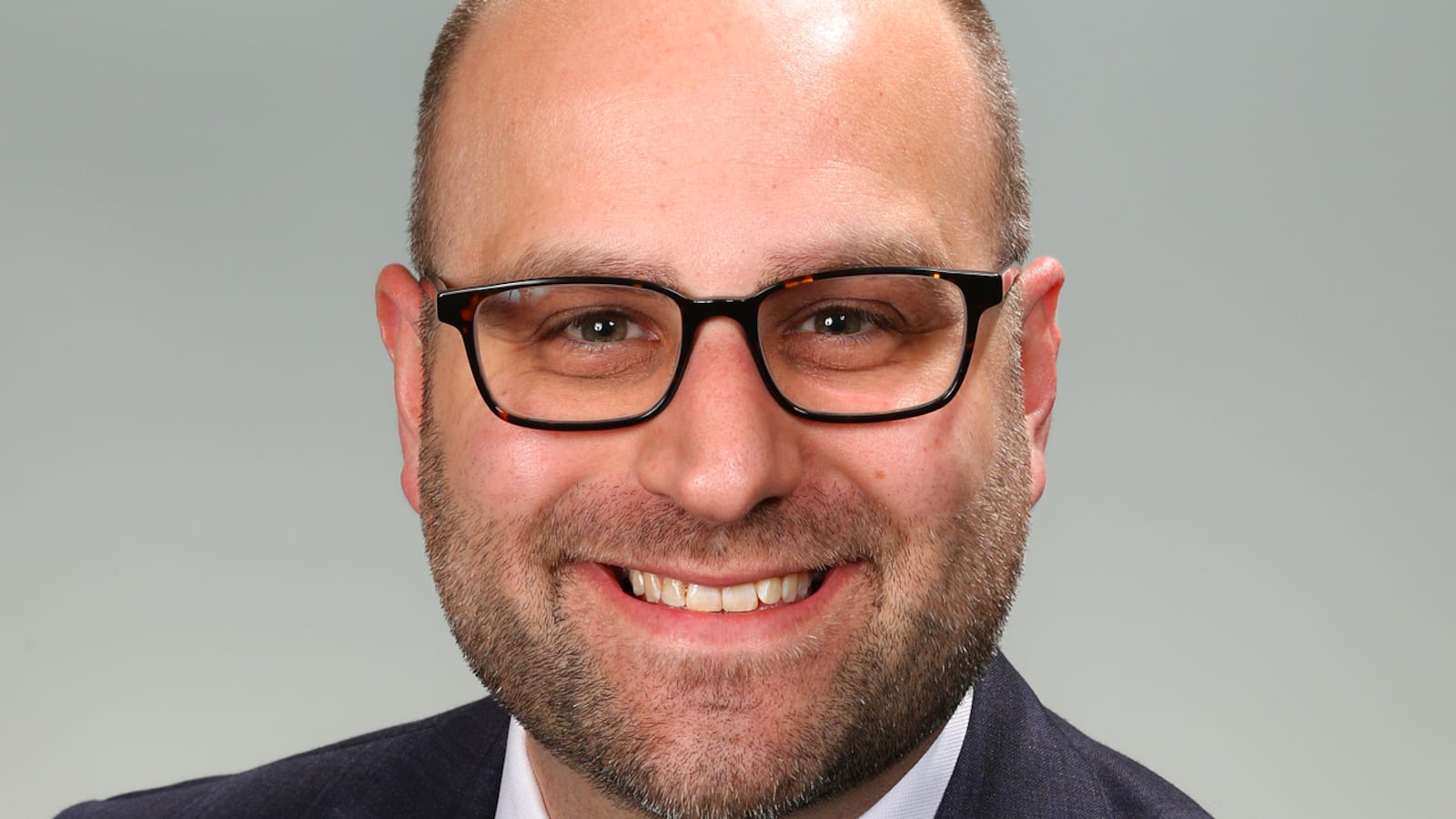 A man with black-framed glasses wearing a dark suit with a yellow tie smiles for a professional headshot.