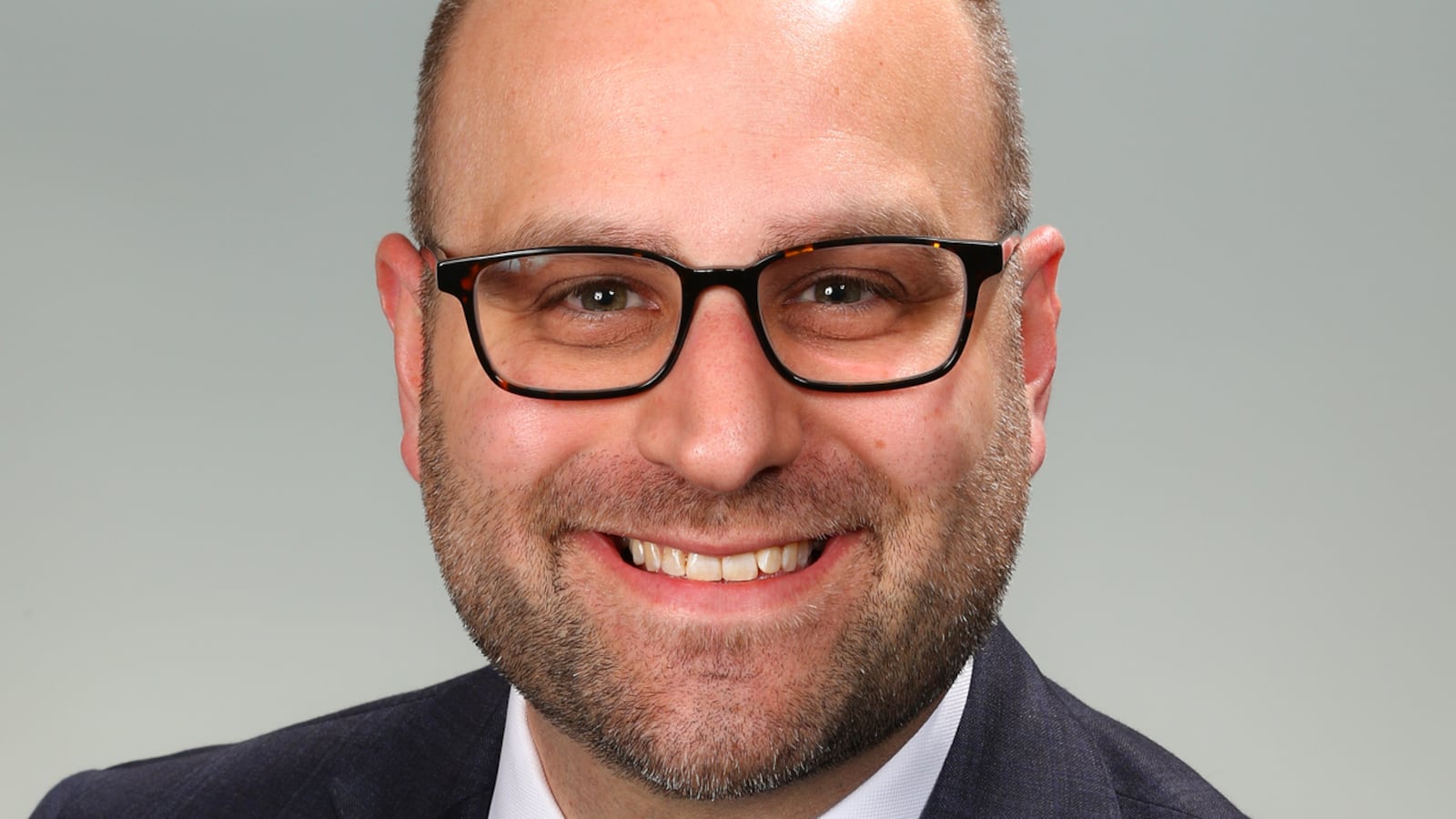 A man with black-framed glasses wearing a dark suit with a yellow tie smiles for a professional headshot.