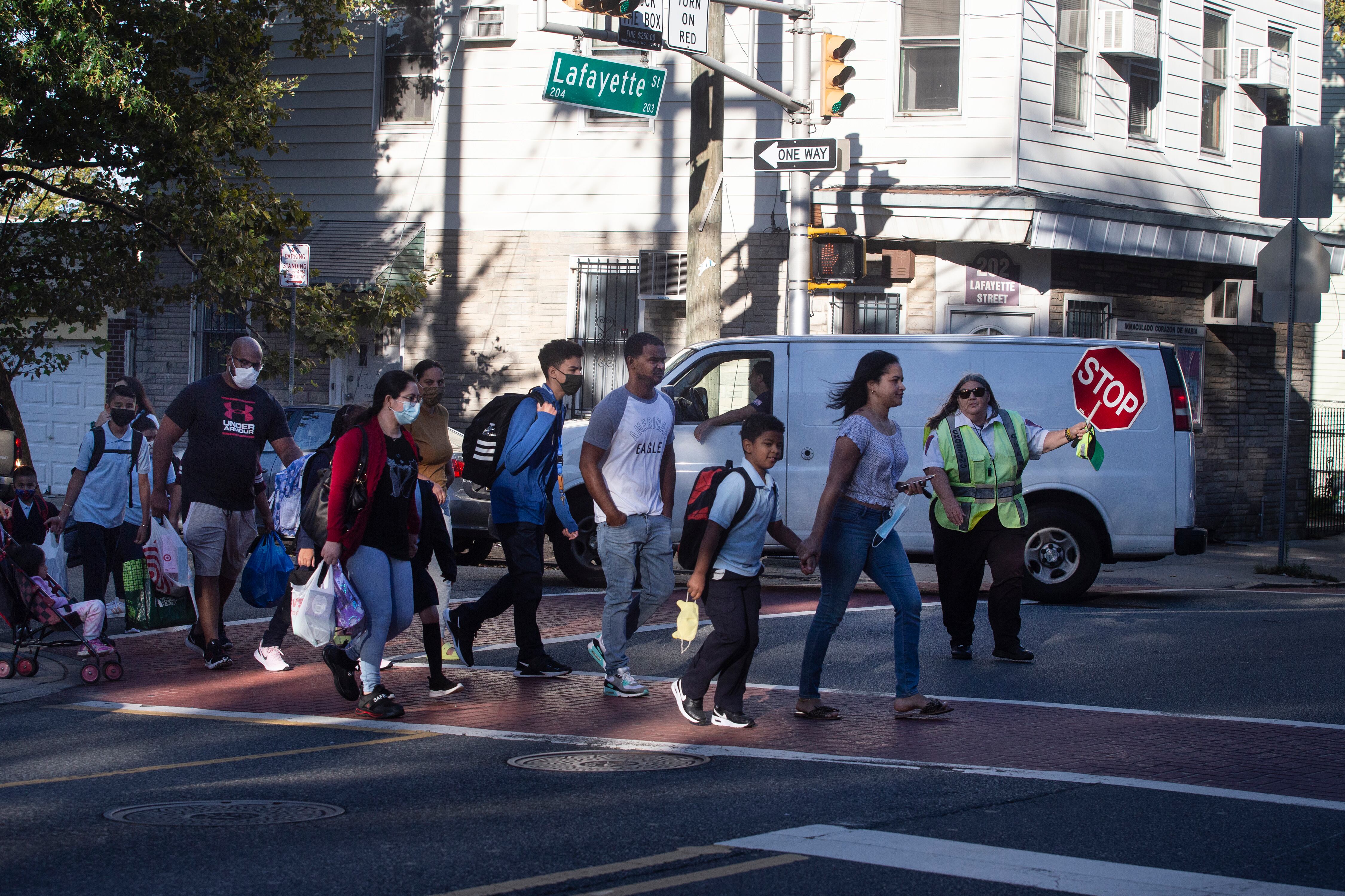 Students wearing backpacks and uniforms cross the street as a crossing guard holds a red stop sign to cars on the street.