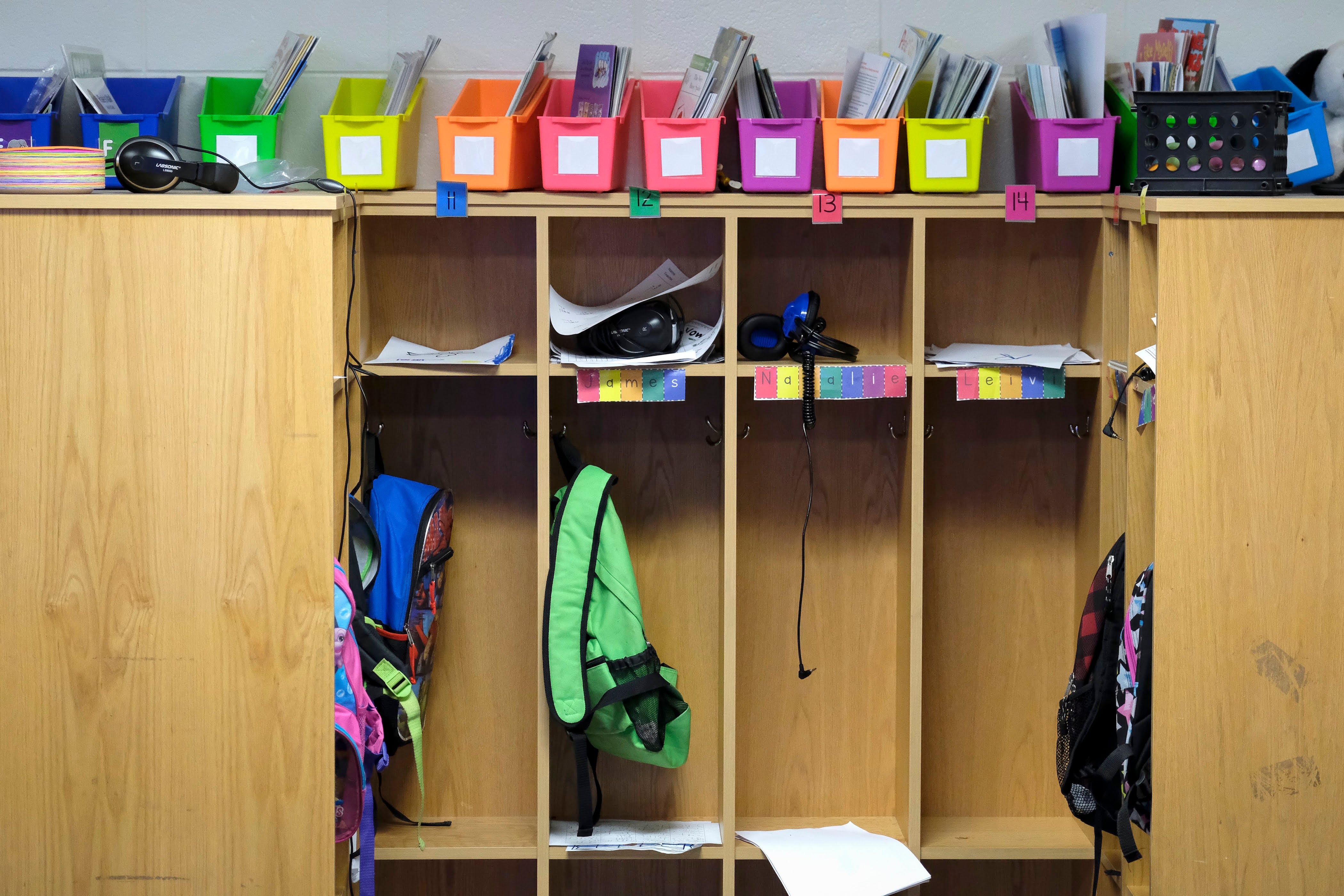 A set of cubbies in a classroom with a couple of backpacks hanging and a row of colorful organizers at the top.