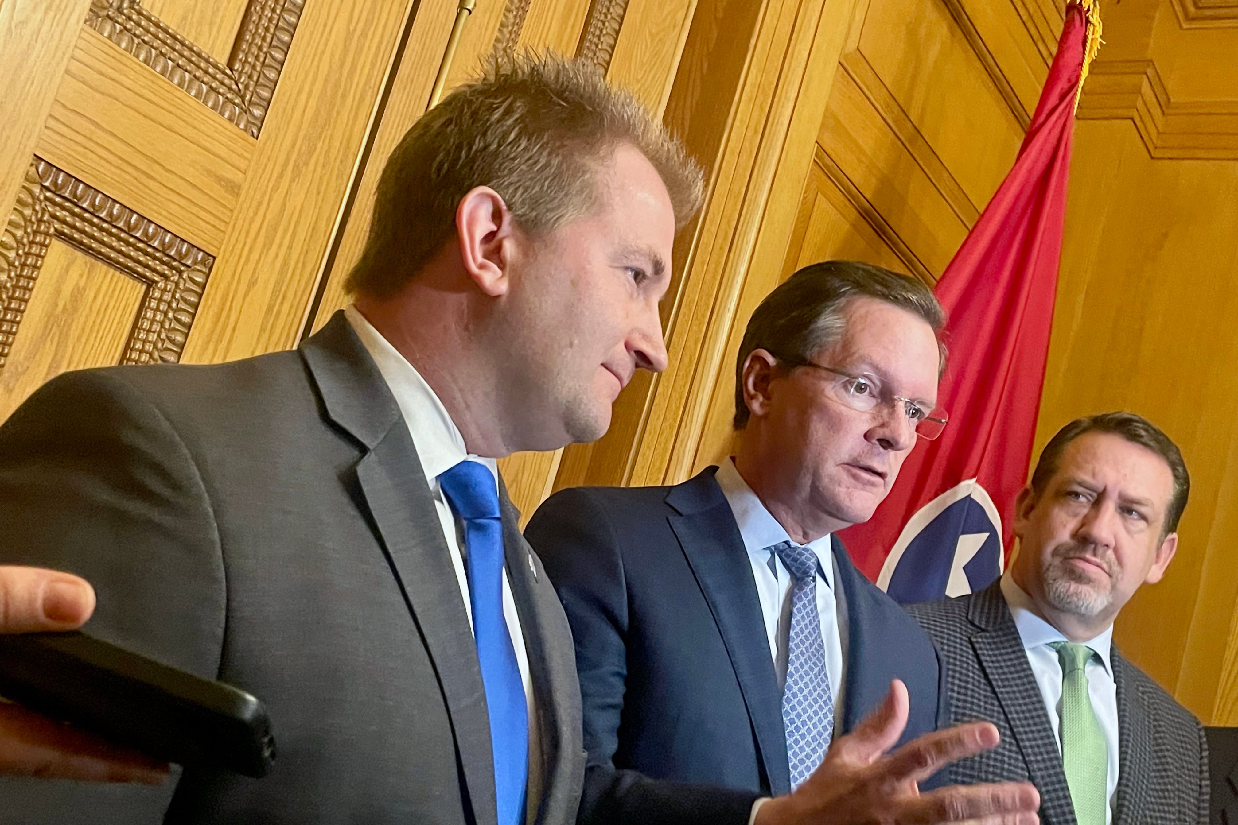 Three men in suits speak in front of a wooden wall with a flag in the background.