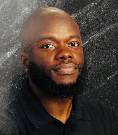 A photograph of a Black man with a shaved head and a full bear wearing a black shirt posing for a school portrait against a grey backdrop.
