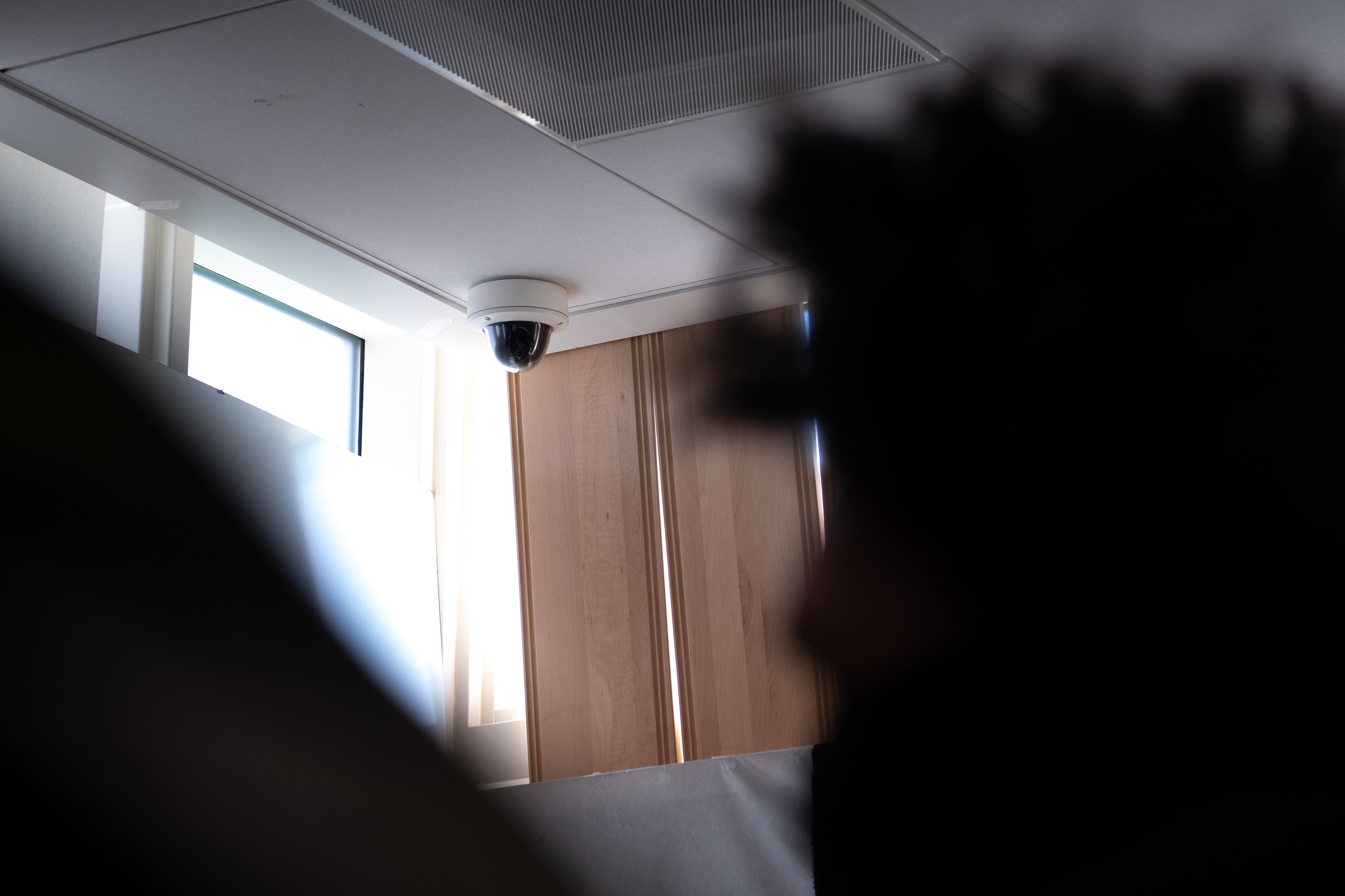 A security camera hangs in the corner of a room, as the silhouette of a student sits in the foreground.