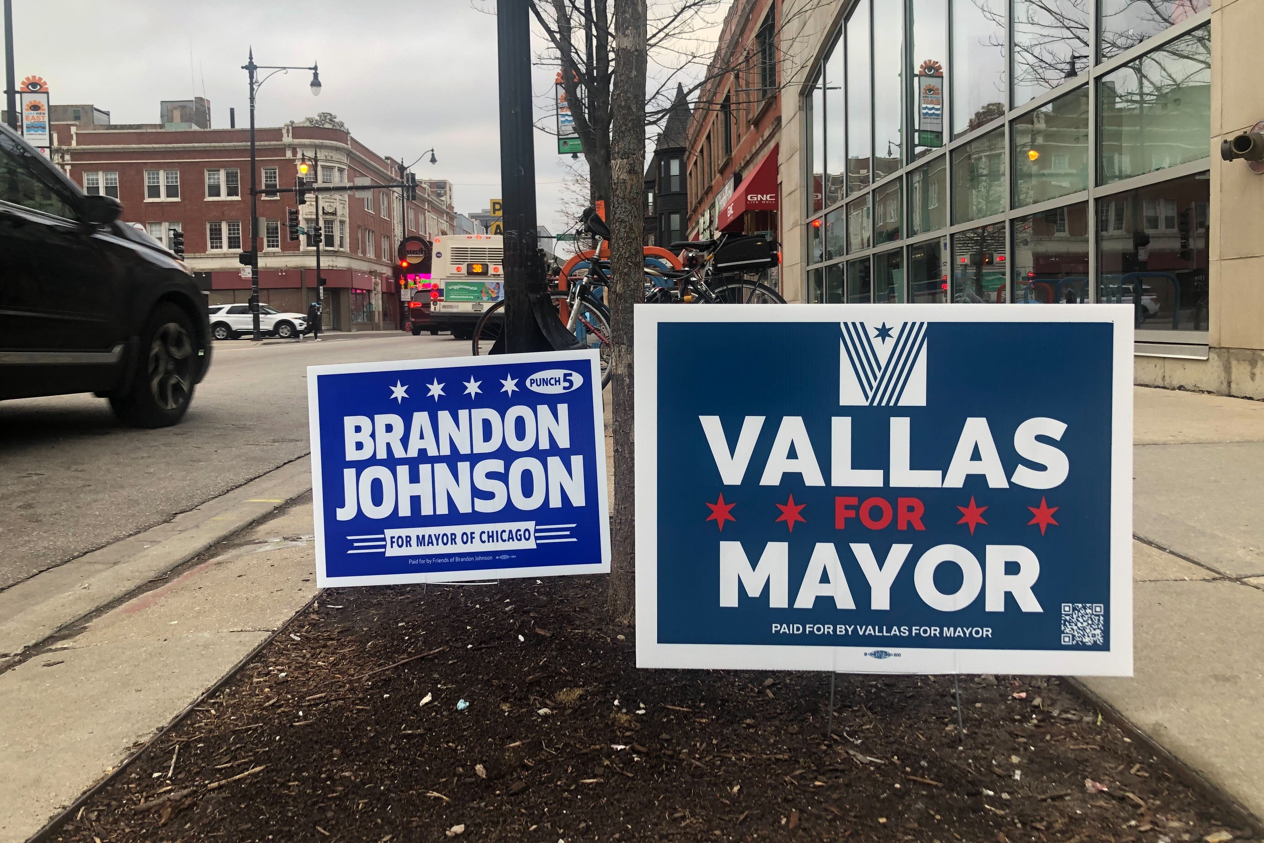 Two campaign yard signs. One for Brandon Johnson and one for Paul Vallas are posted along a Chicago sidewalk.
