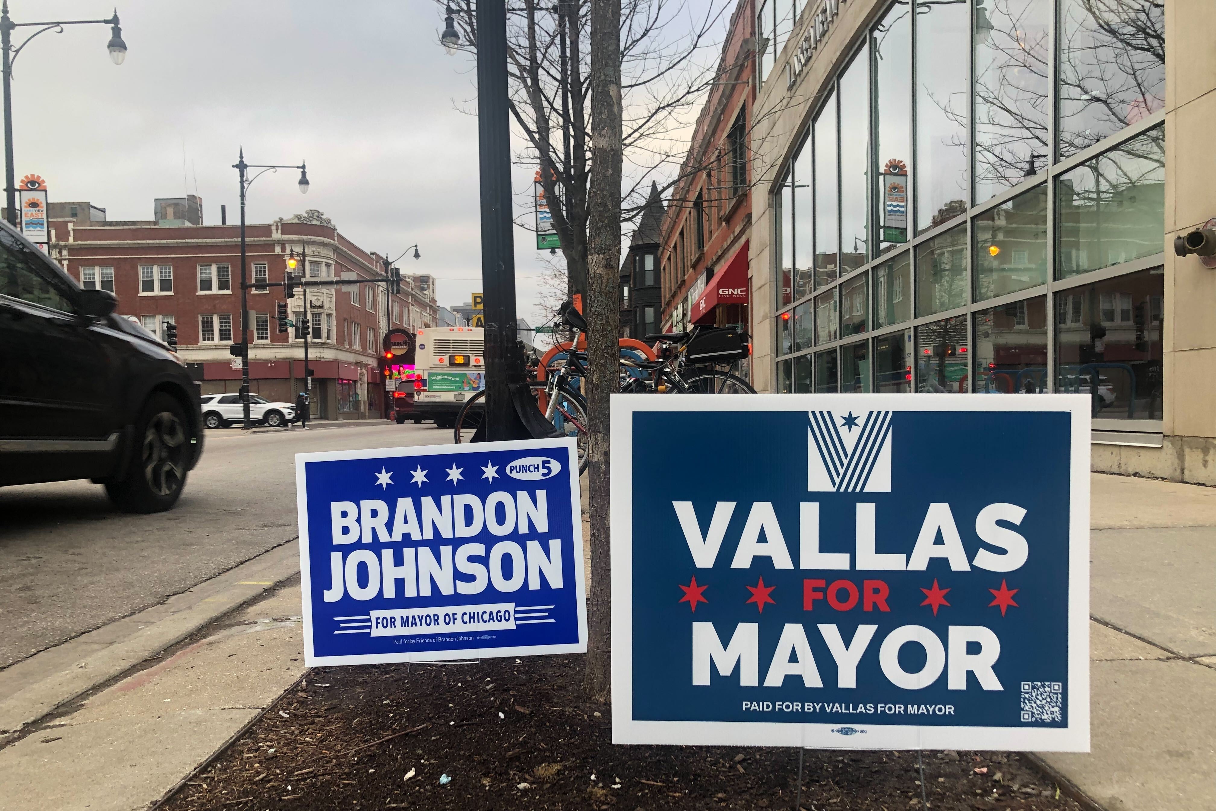 Two campaign yard signs. One for Brandon Johnson and one for Paul Vallas are posted along a Chicago sidewalk.