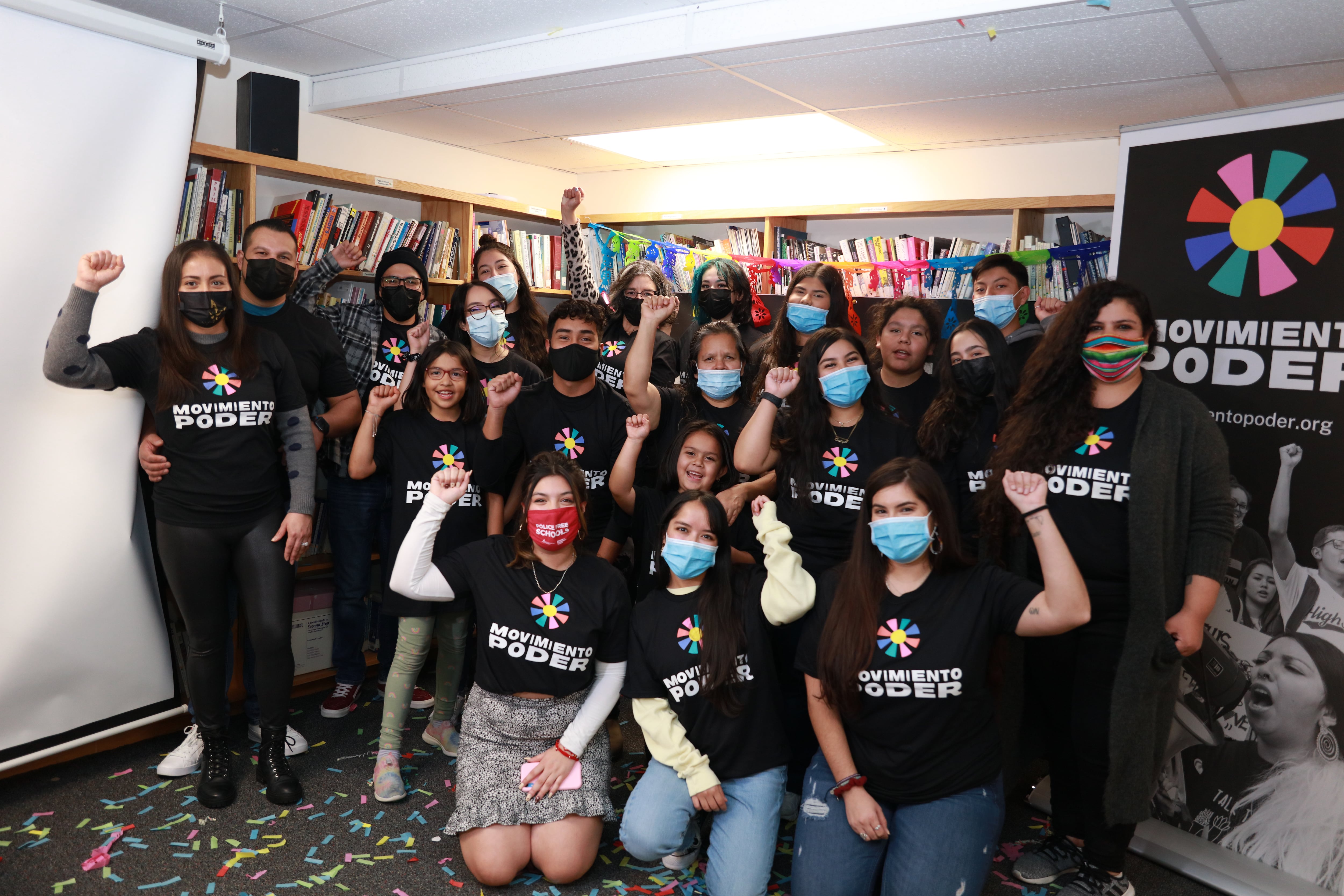 A group of people wearing Movimiento Poder T-shirts pose for a photo. Many of them have a fist raised in the air. They are wearing face masks.