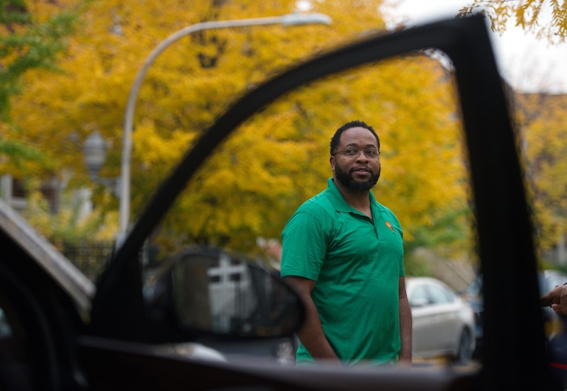 A man wearing a green shirt poses for a photograph outside with a car window in the foreground and yellow trees in the background.