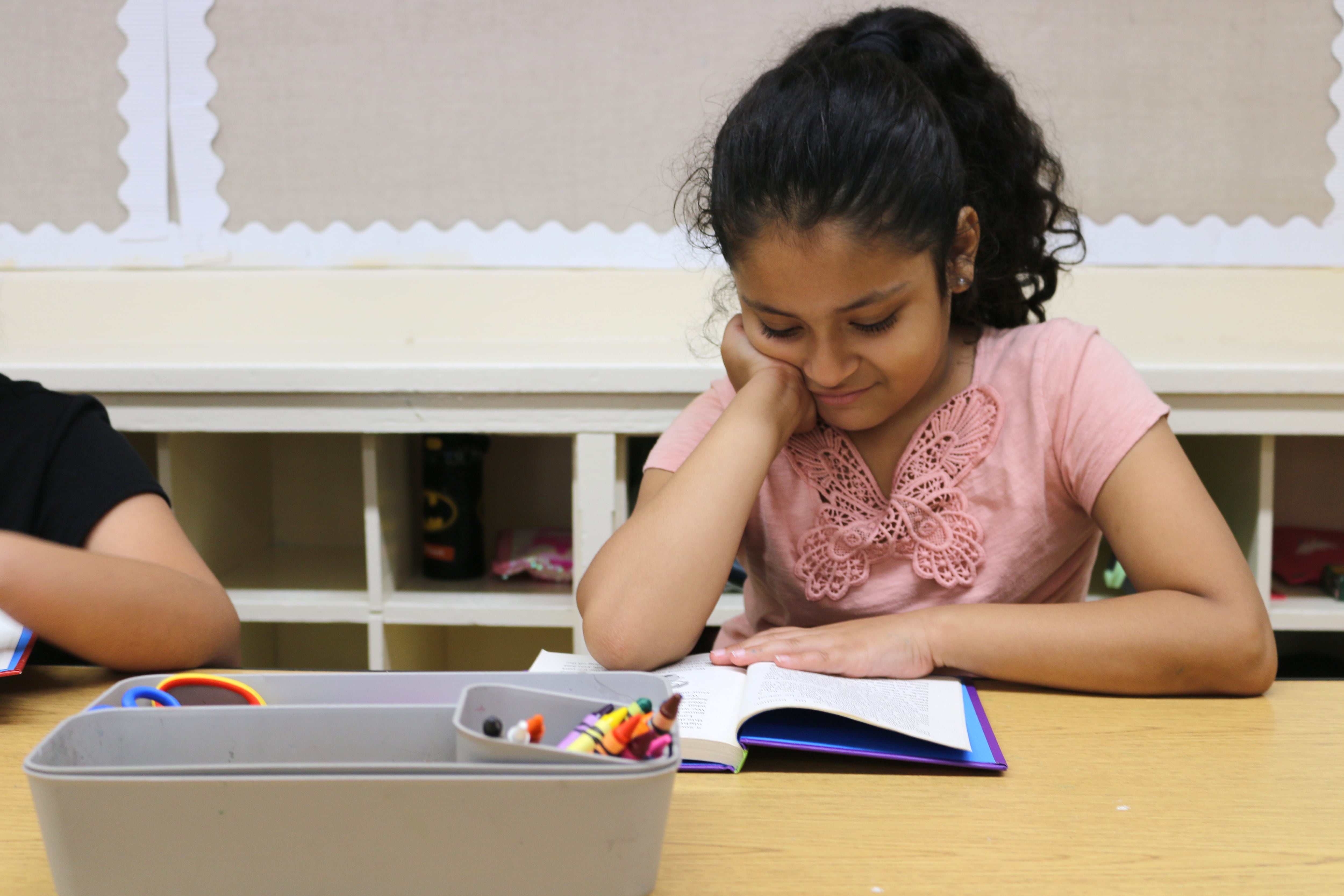 A young girl in a peach top reads a book at a table.