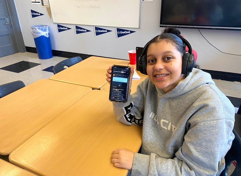 A person wearing a grey hoodie and headphones holds out a phone with a wooden desk in the background and a string of blue banners on the back wall.