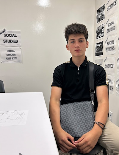 A high school student sits at a white table with a white wall in the background.
