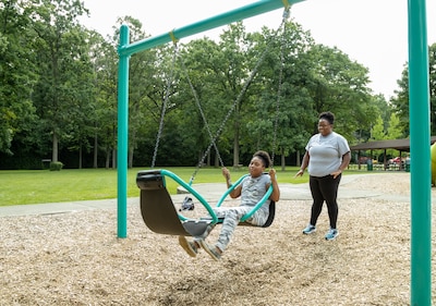 A child on the right in a swing while an adult stands behind her both wearing light colored t-shirts outside with green trees in the background.