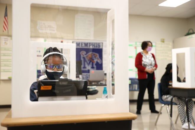 A fifth grade girl wearing a mask works on a laptop behind a clear desktop barrier. A woman stands in the background near another girl working at a desk behind a partition.