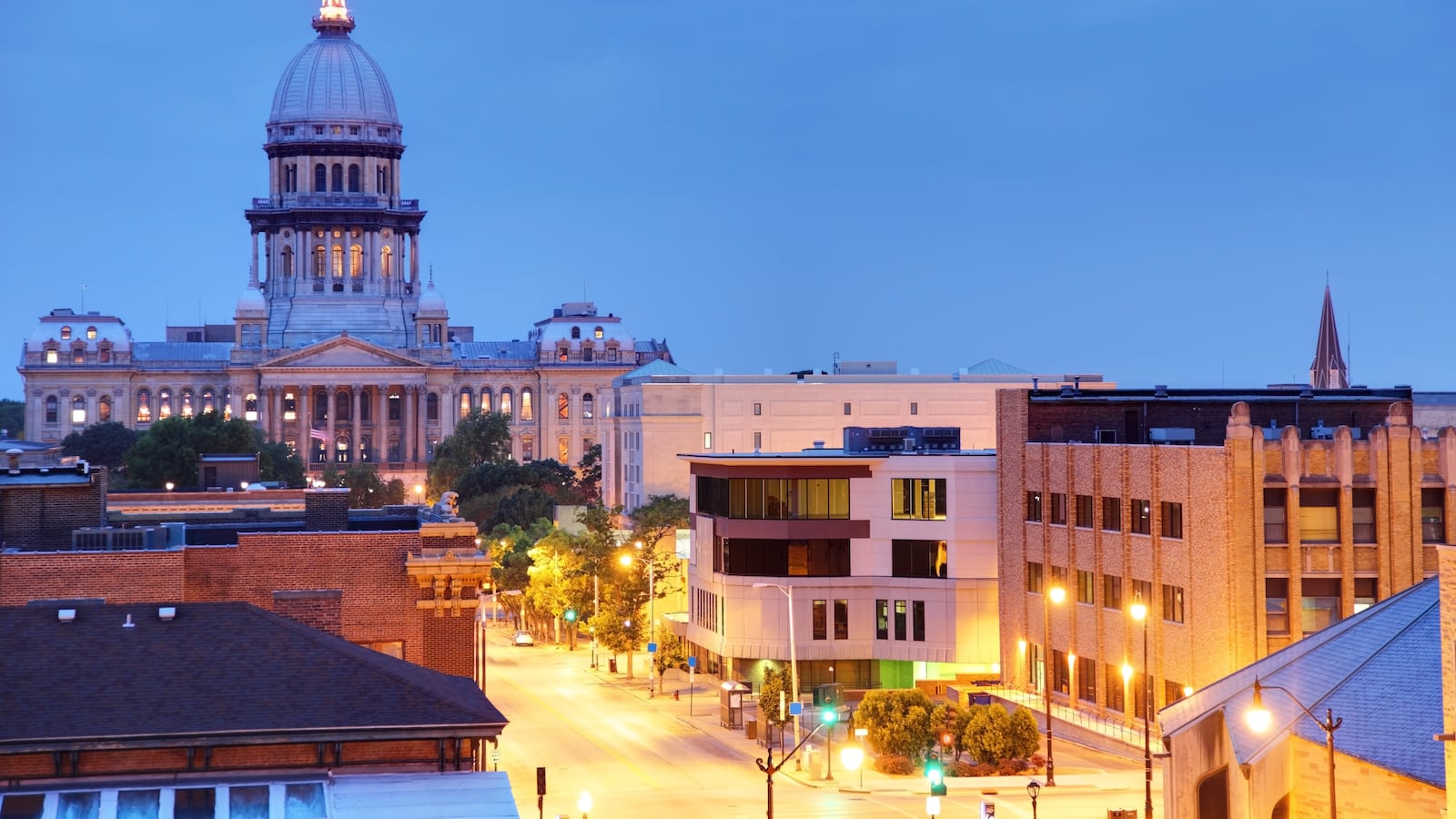 Springfield, IL capitol sits in the background of the photo, barely lit as the sky is dark blue. Buildings lining the road leading to the capitol building are brightly lit in the foreground of the photo.