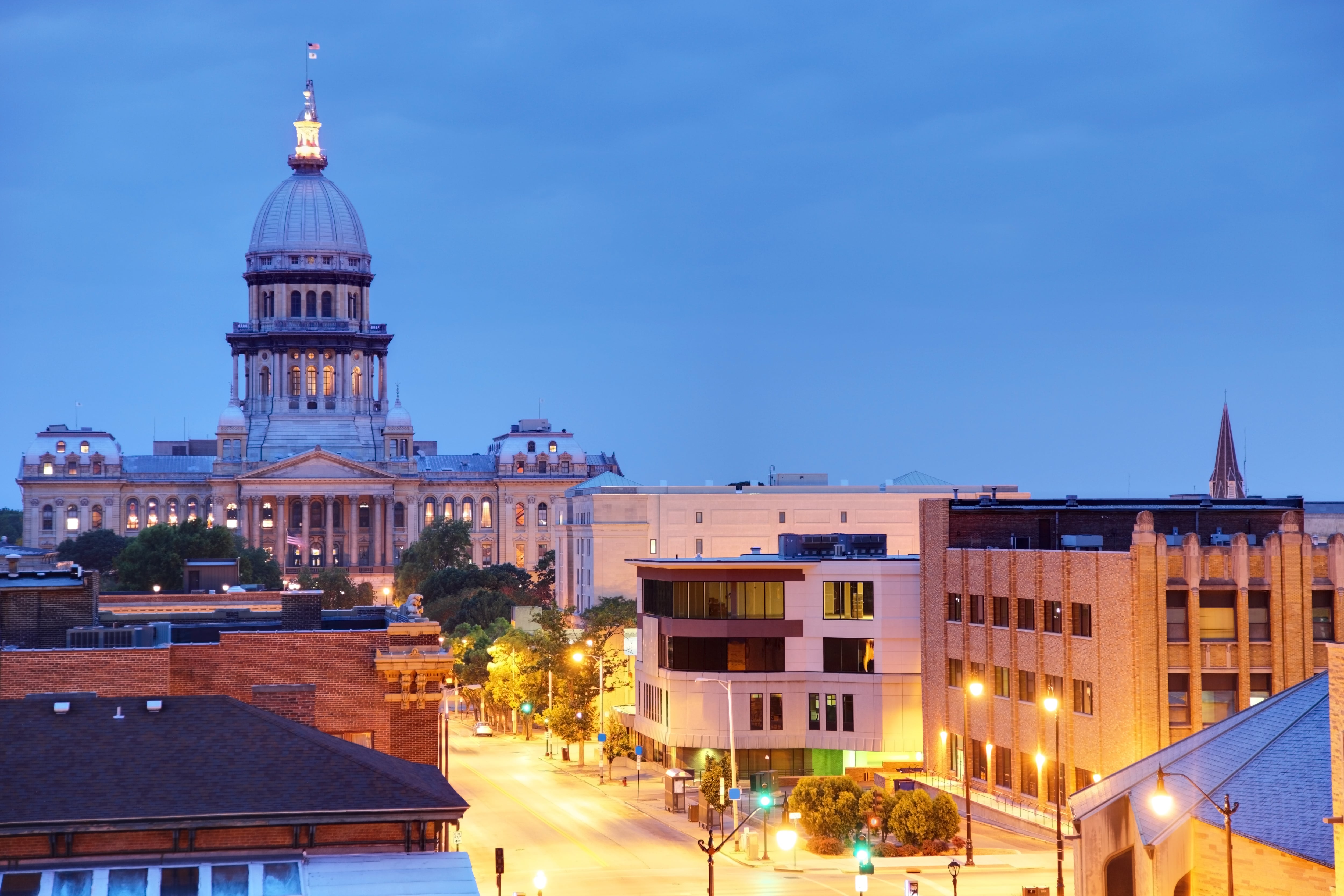 Springfield, IL capitol sits in the background of the photo, barely lit as the sky is dark blue. Buildings lining the road leading to the capitol building are brightly lit in the foreground of the photo.