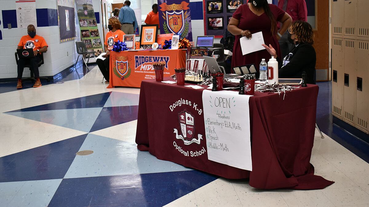 A maroon table for Douglass Optional School and Ridgeway Middle sit in a school hallway with blue and white tile. The desks are staffed by two people each, wearing their school’s colors.
