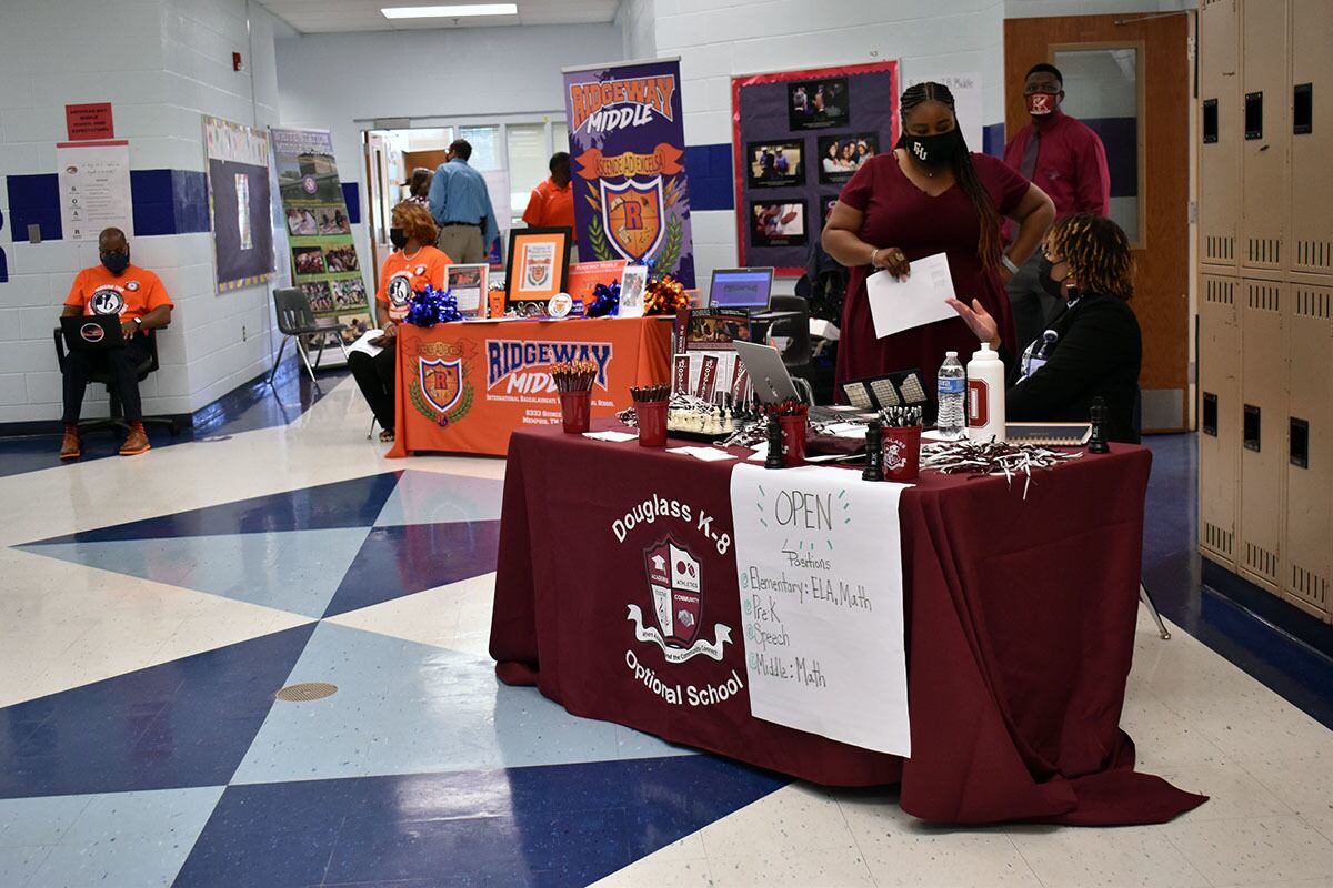 A maroon table for Douglass Optional School and Ridgeway Middle sit in a school hallway with blue and white tile. The desks are staffed by two people each, wearing their school’s colors.