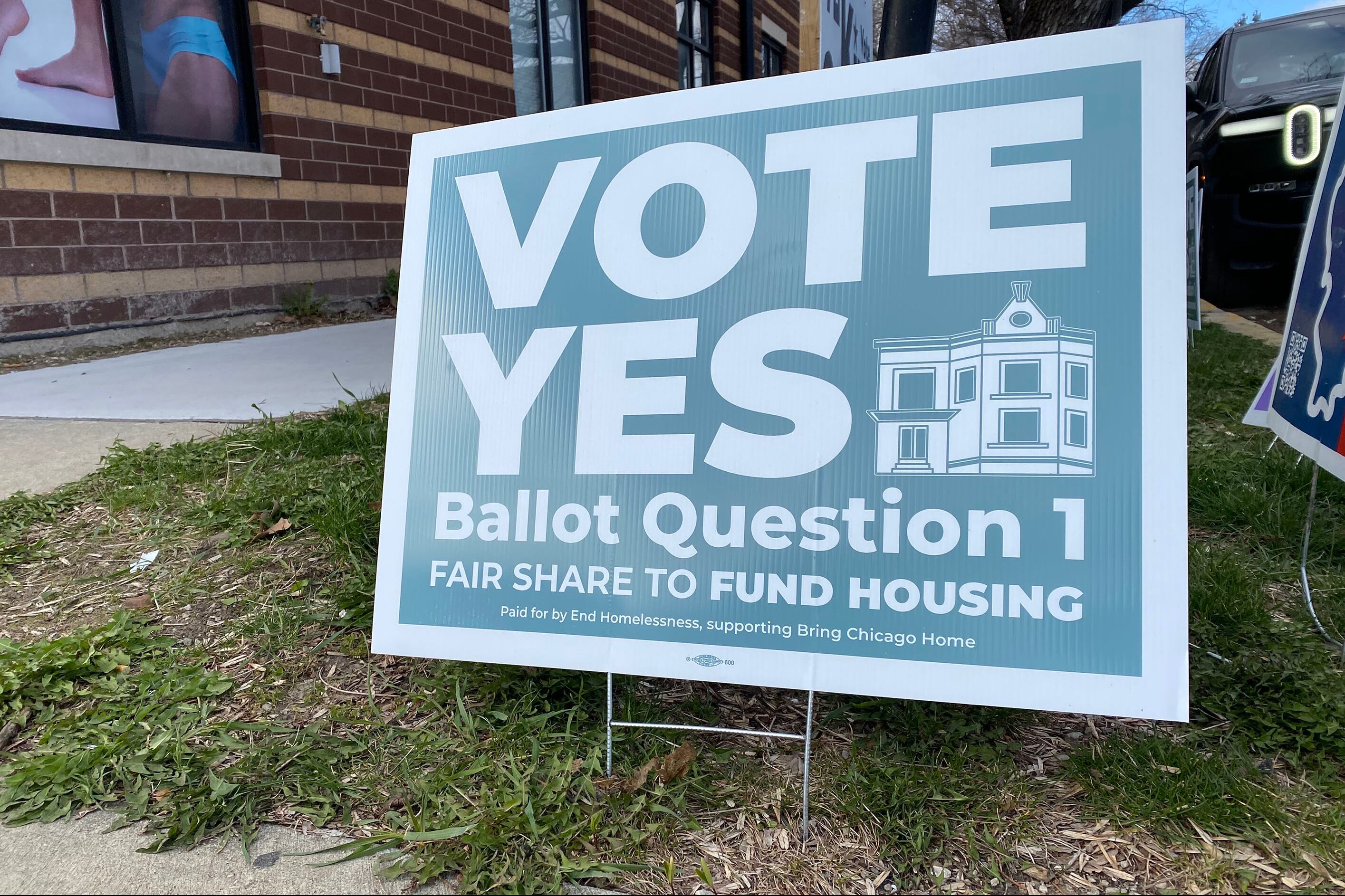 A blue and white sign sits in a grassy lawn outside of a red brick building. Some of the words on the sign read "Vote Yes Ballot Question 1."