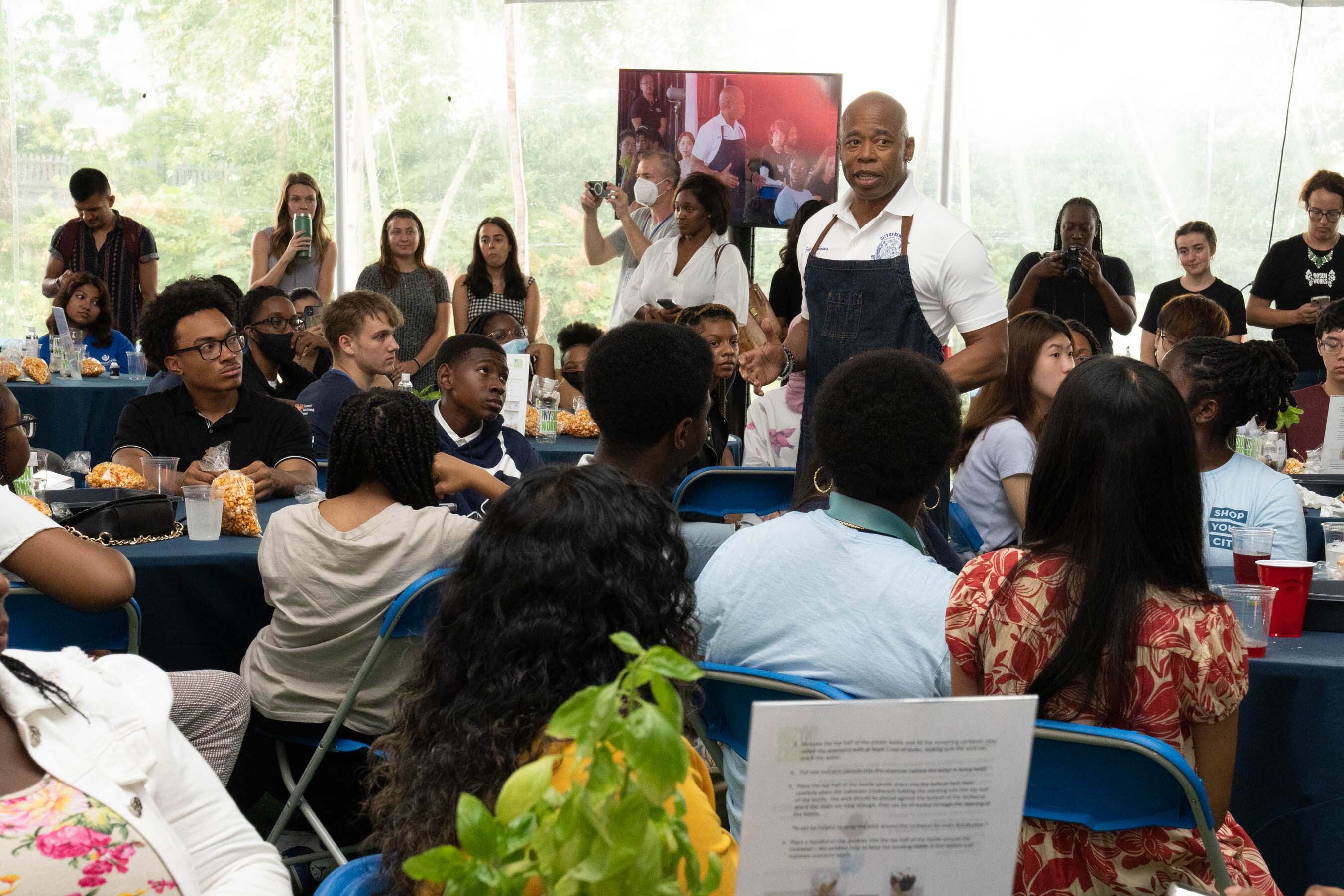 A man in an apron and white shirt stands in the middle of a crowd of people sitting in chairs.