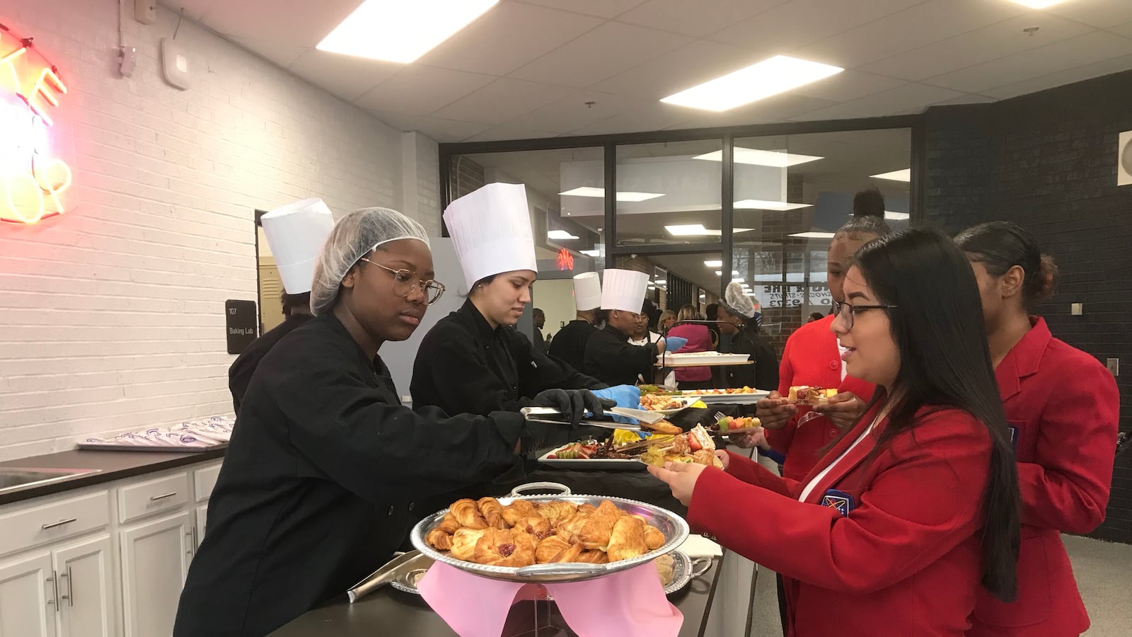 A student wearing a hair net and two wearing chefs hats serve people in line at a buffet table. 