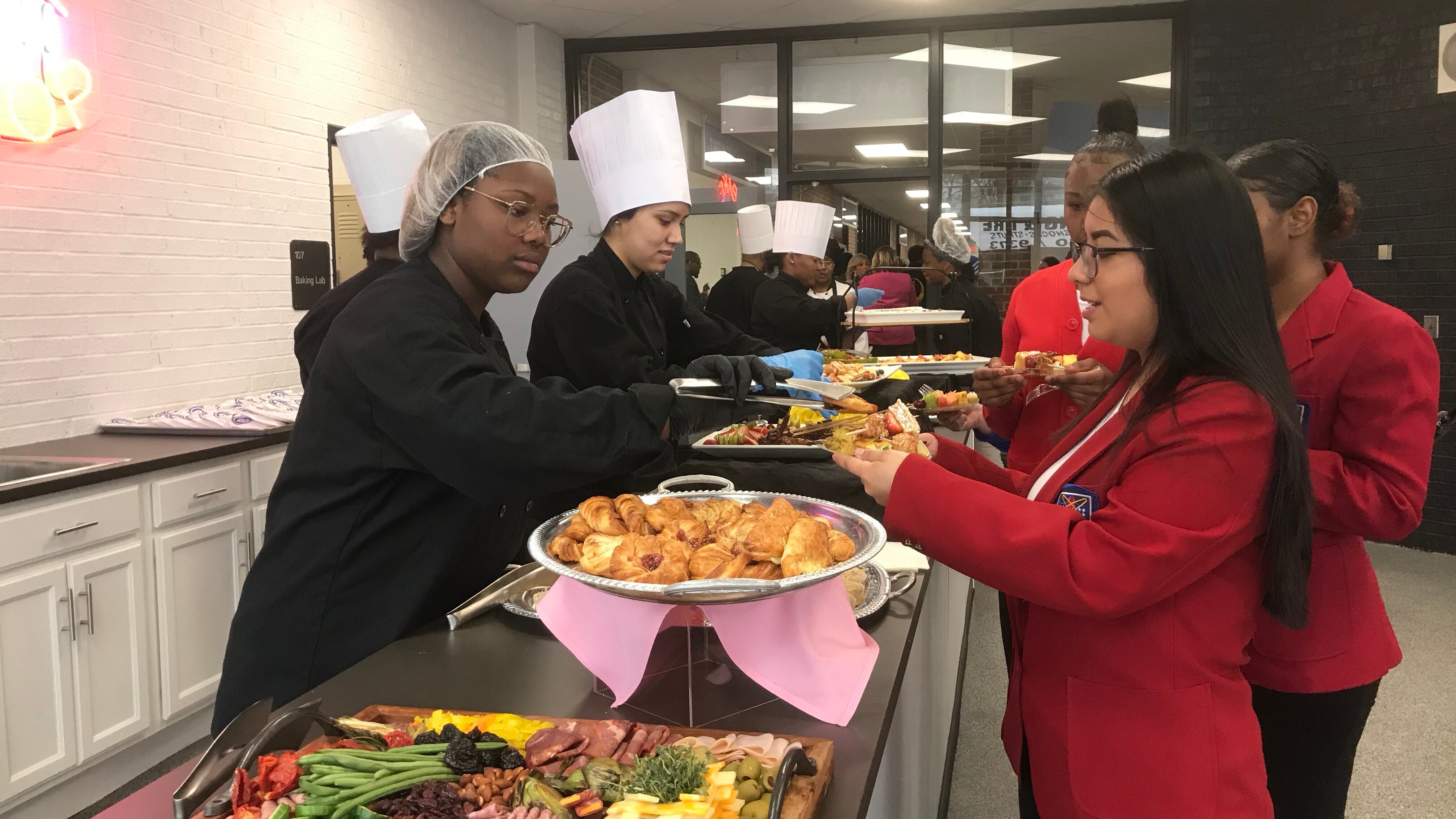 A student wearing a hair net and two wearing chefs hats serve people in line at a buffet table. 