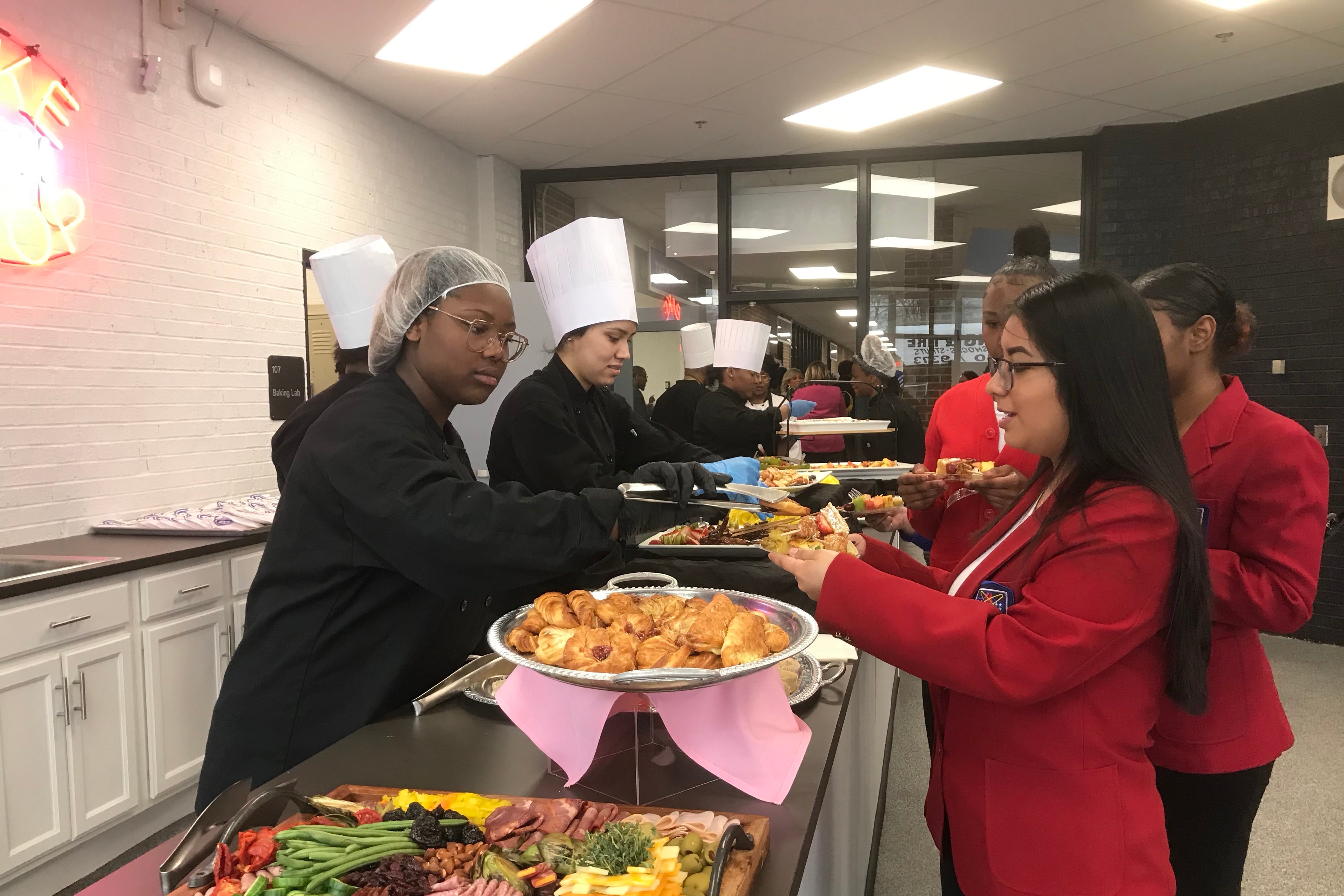 A student wearing a hair net and two wearing chefs hats serve people in line at a buffet table.