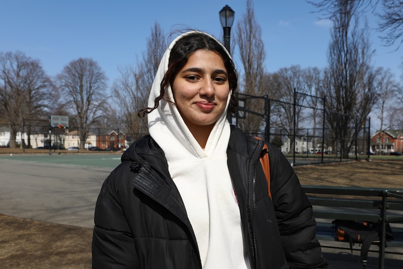 A high school student wearing a white hoodie and a black jacket smiles for a portrait outside on a cold and sunny day.