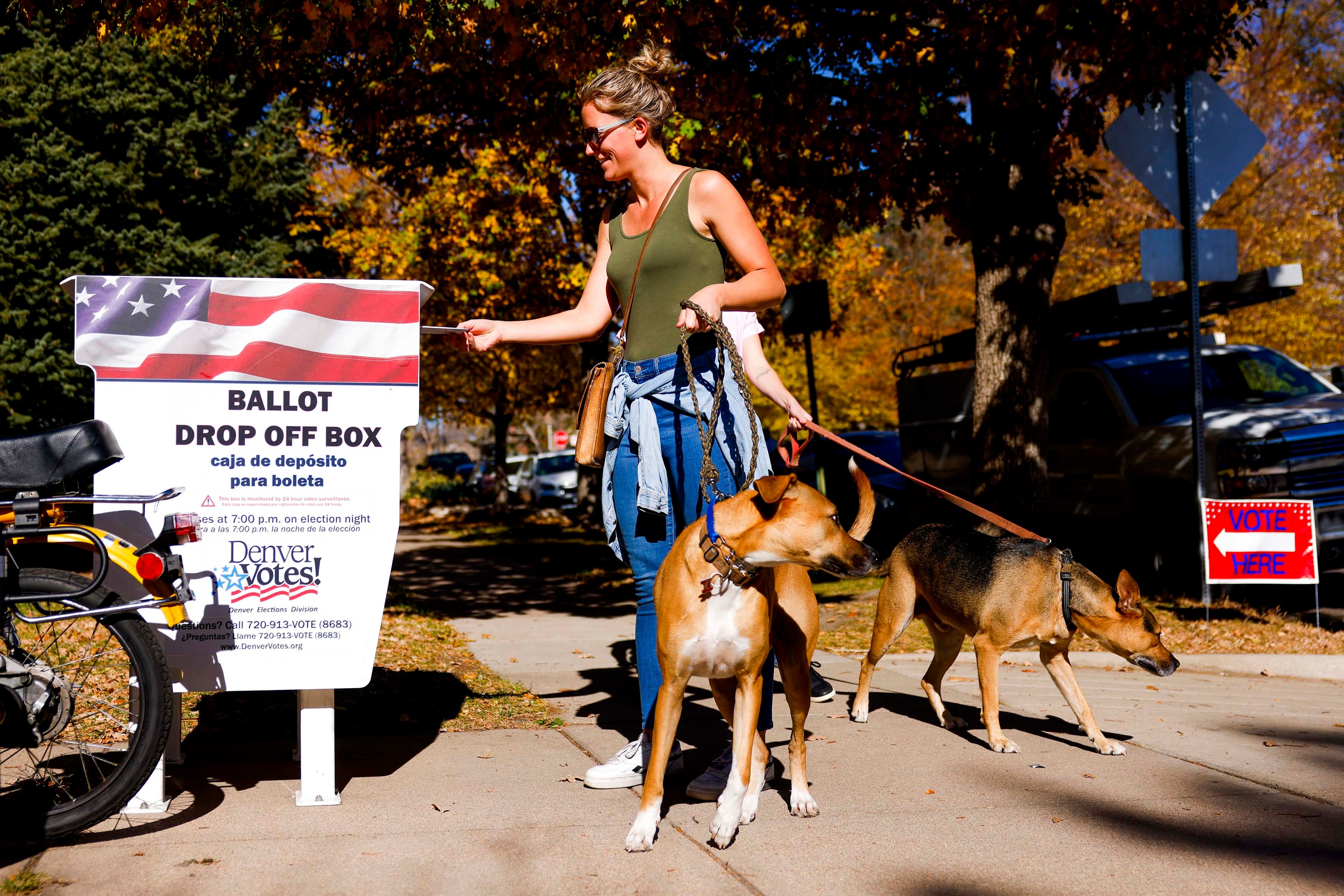 A smiling woman holding two dogs on leashes places her ballot in a drop box outside the La Familia Recreation Center in Denver.