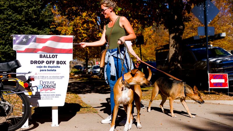 A smiling woman holding two dogs on leashes places her ballot in a drop box outside the La Familia Recreation Center in Denver.