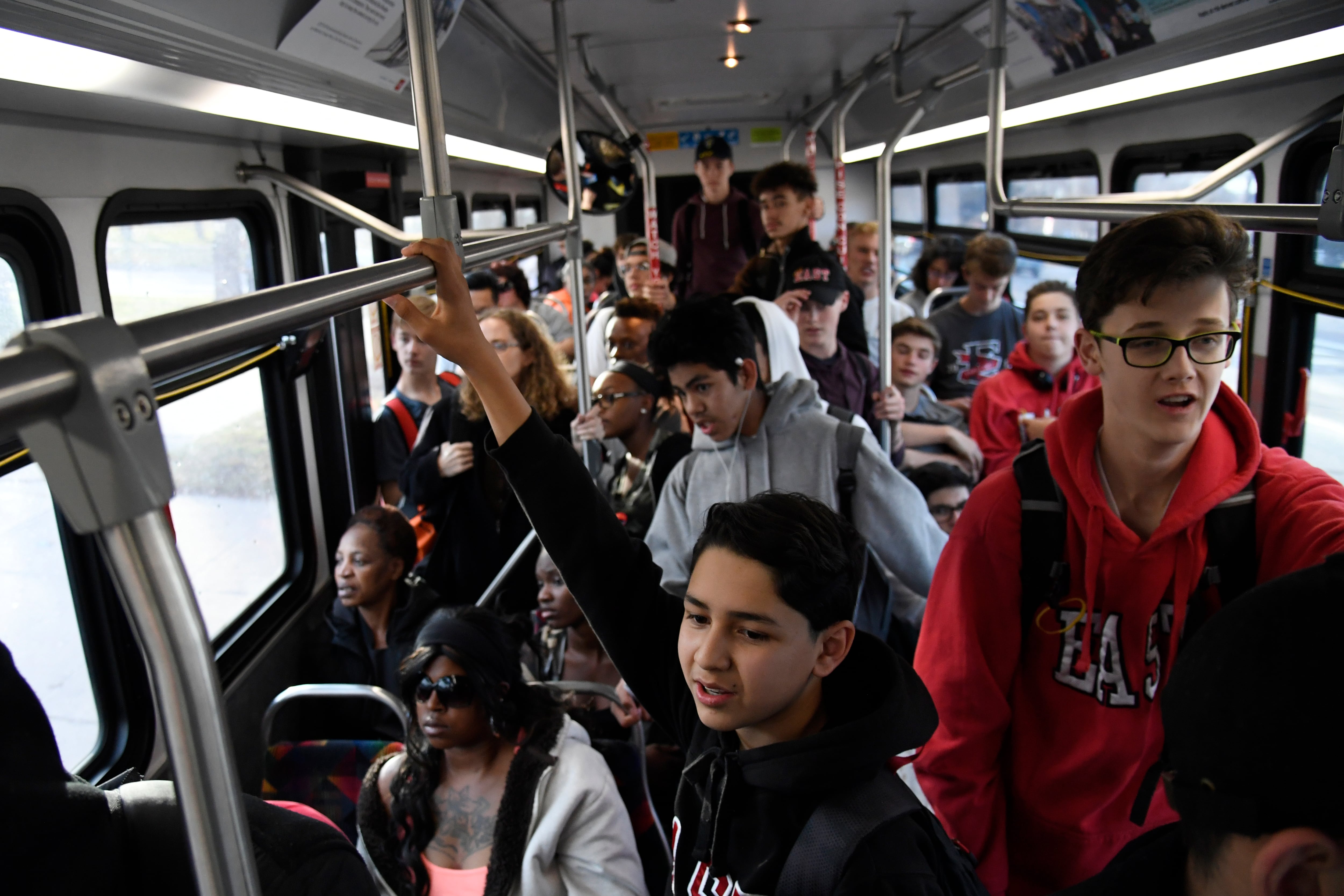 A city bus is packed with teenagers, many of them wearing hoodies, hanging from the hand rails.