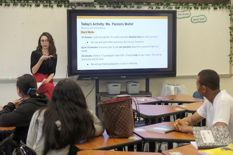 A photograph of a white woman teacher standing at the front of the room by a dry erase board with three high school students sitting at their desks in a classrom.