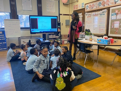 A teacher with medium length dark hair and wearing a colorful cardigan, stands next to a class of young students all sitting on a blue rug. There is a projector, a desk and posters in the background.