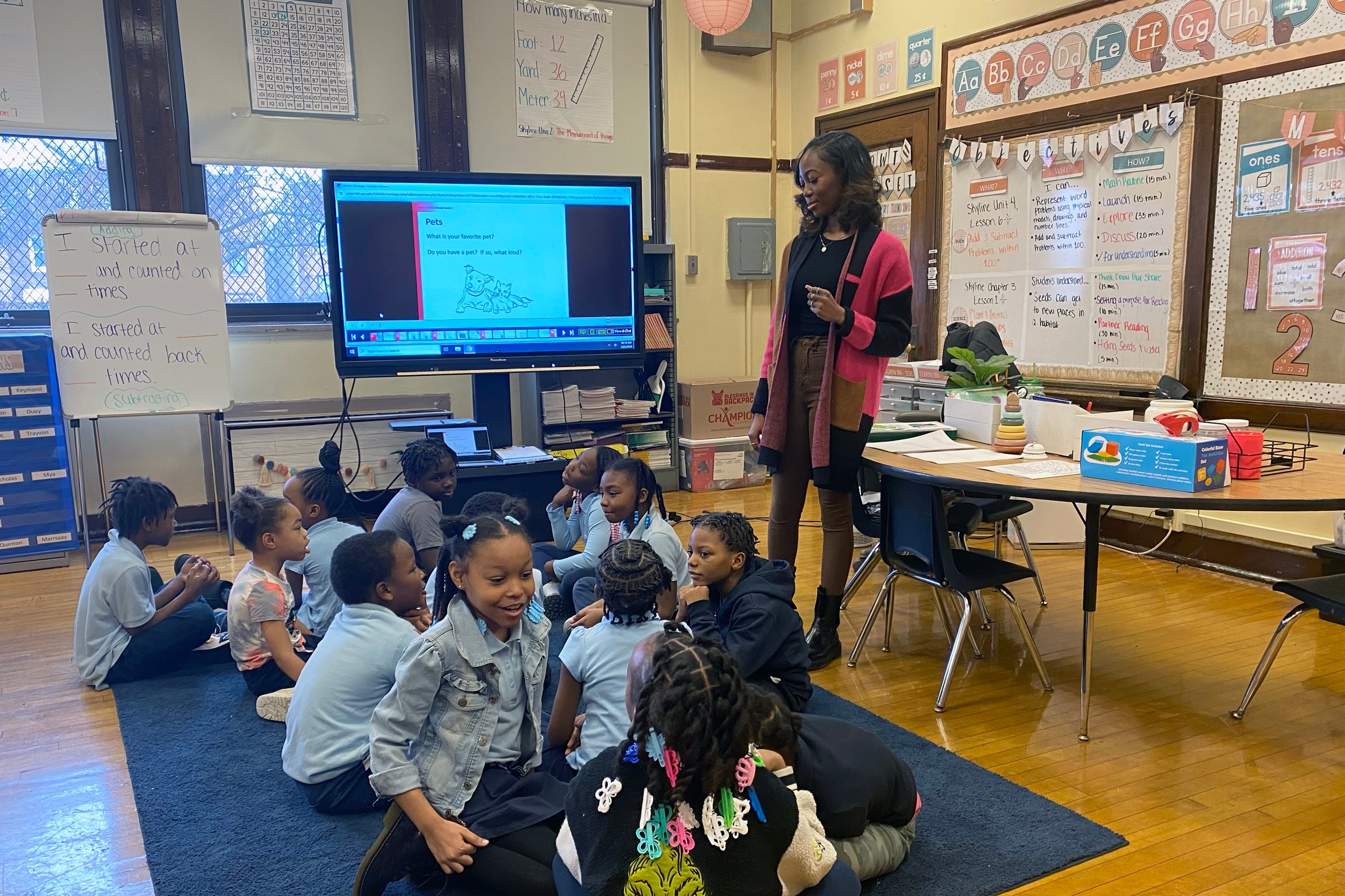 A teacher with medium length dark hair and wearing a colorful cardigan, stands next to a class of young students all sitting on a blue rug. There is a projector, a desk and posters in the background.