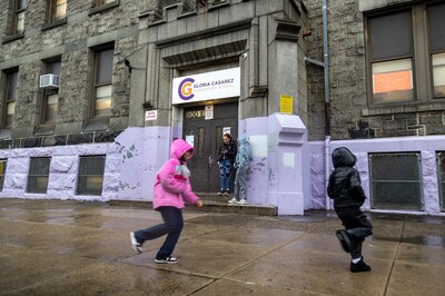 Two children play in the foreground while an adult and a child talk at the front entrance of a school building.