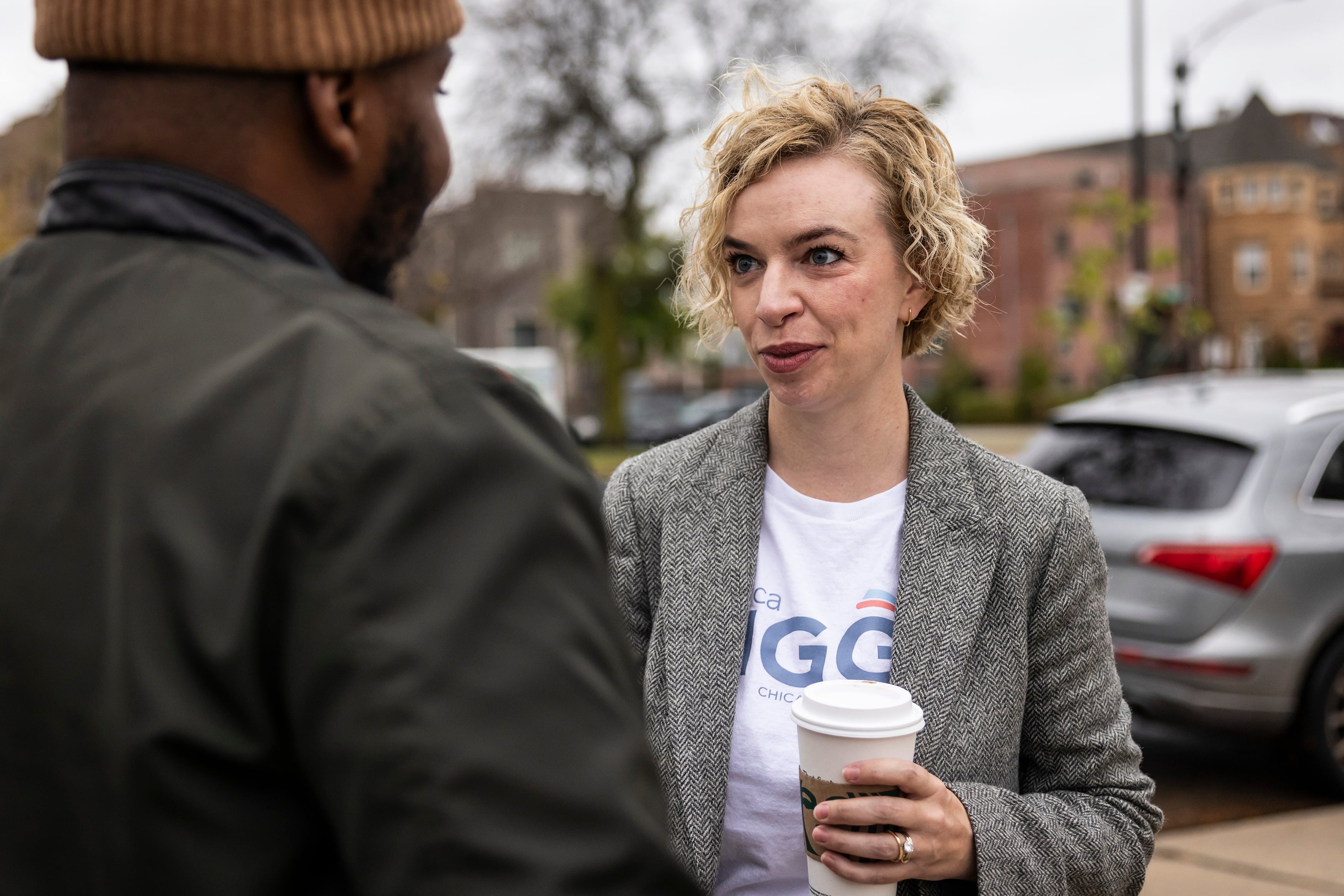 A white woman with short blonde hair wearing a grey suit jacket and is holding a starbucks coffee cup speaks to a Black man in a green jacket and orange beanie outside on a cloudy day.