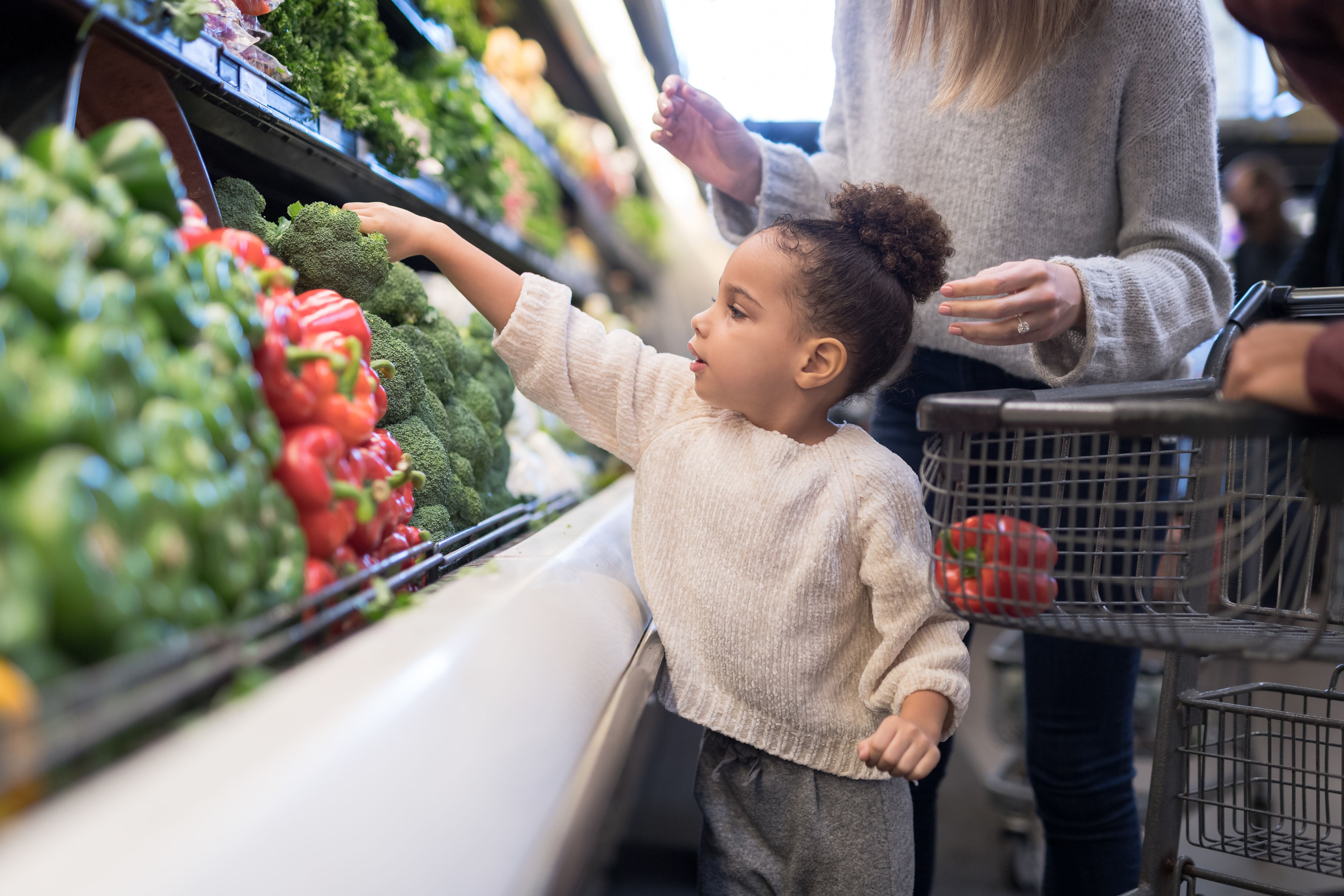 A young child reaches for a head of broccoli in a grocery store with an adult behind them.