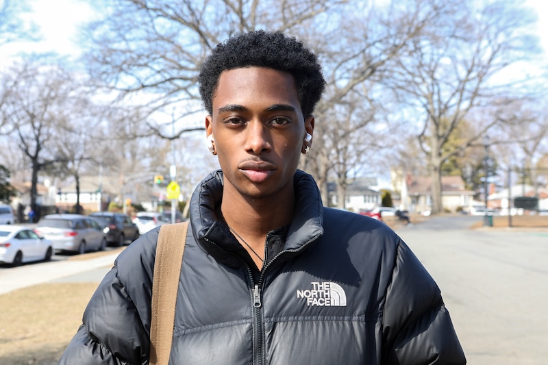 A Black student wearing a black jacket poses for a portrait outside on a sunny, cold day.