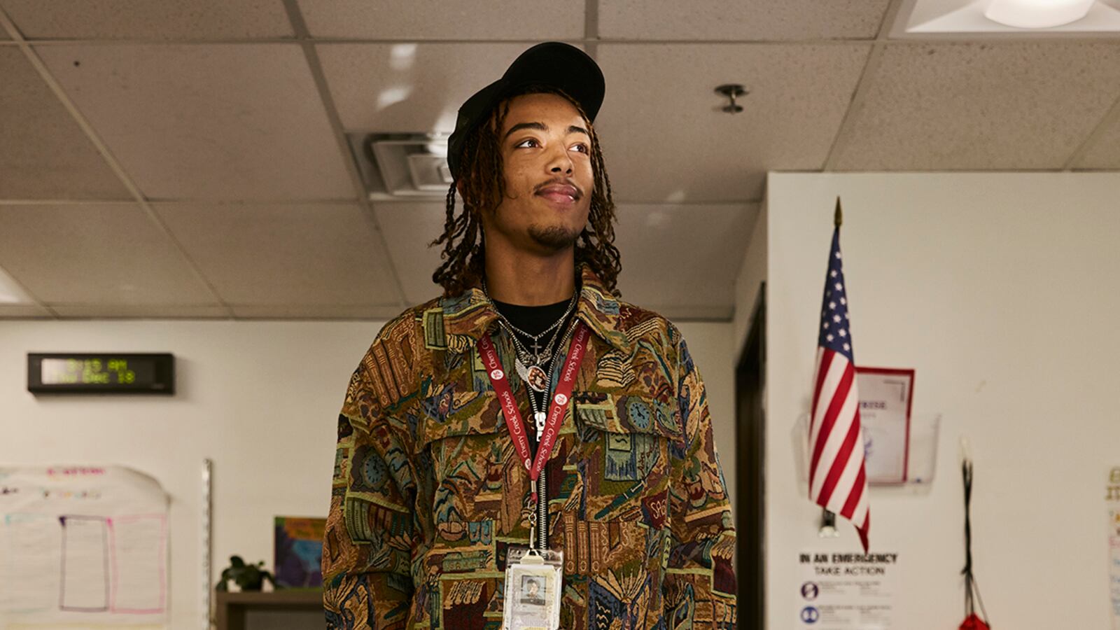 A photograph of a young Black man standing in a classroom posing for a photograph while a ray of light touches his face.