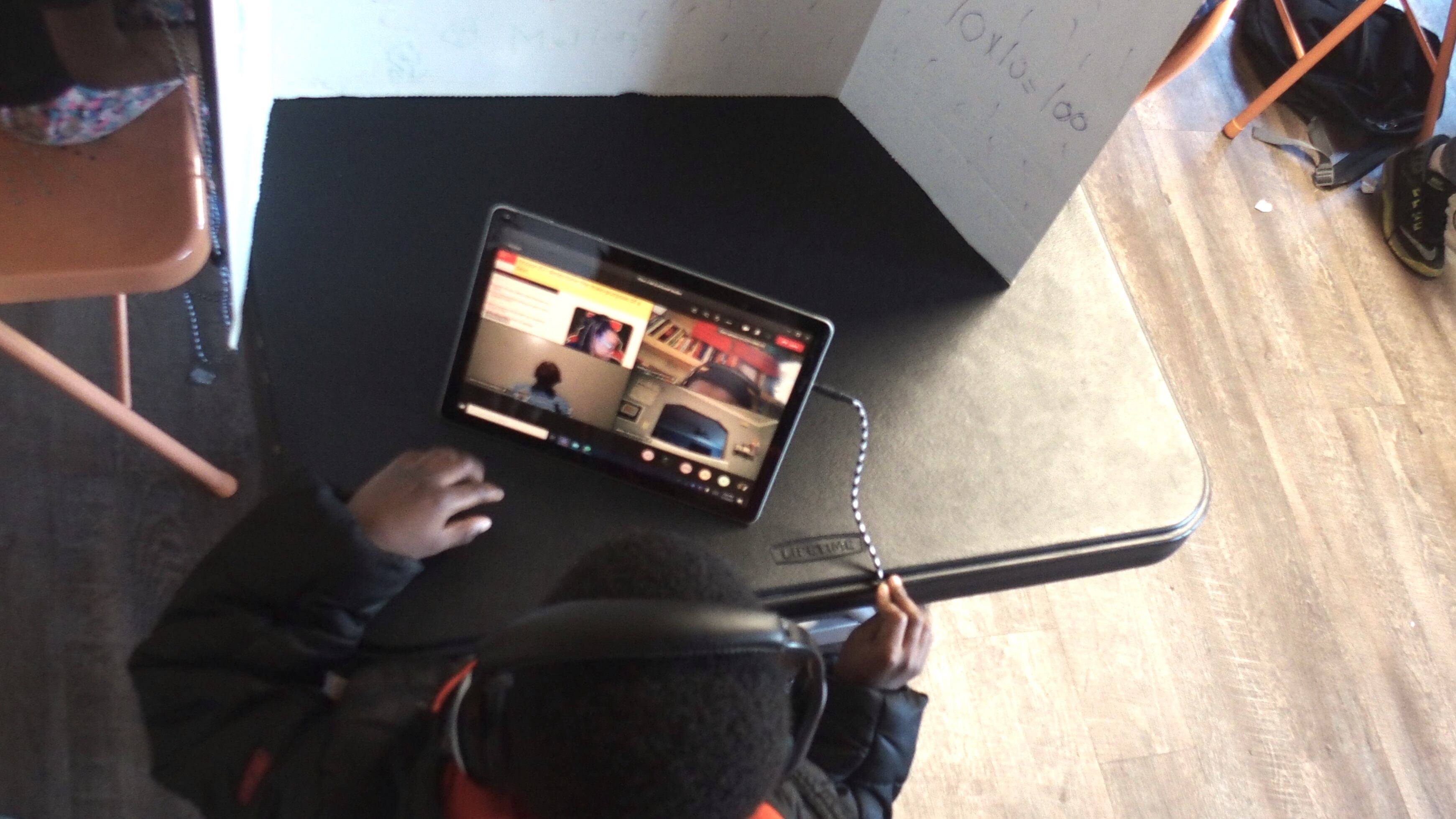 A young boy in a black and orange hoodie conducts virtual learning on his computer, sitting at a black partitioned desk.