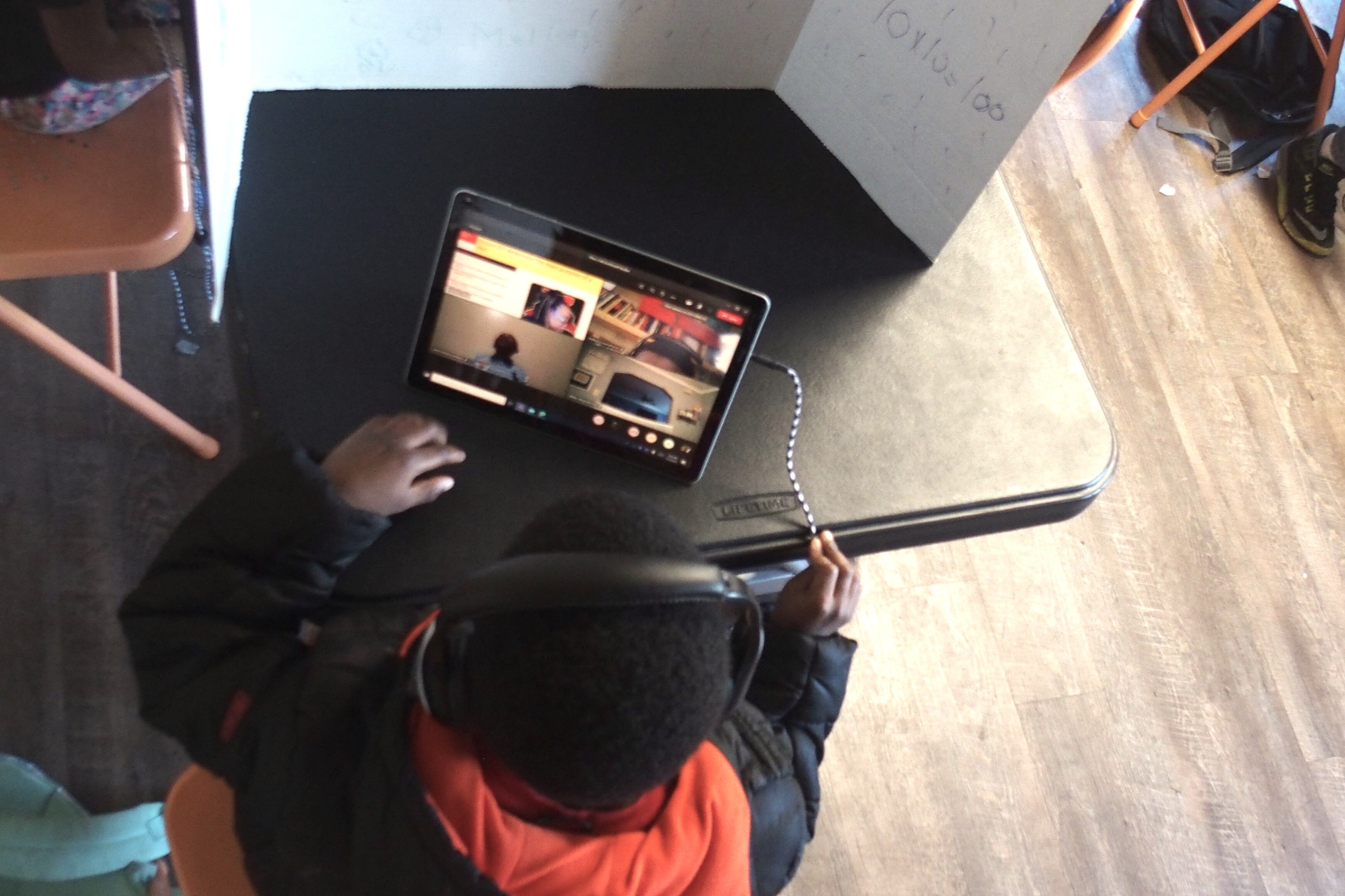 A young boy in a black and orange hoodie conducts virtual learning on his computer, sitting at a black partitioned desk.