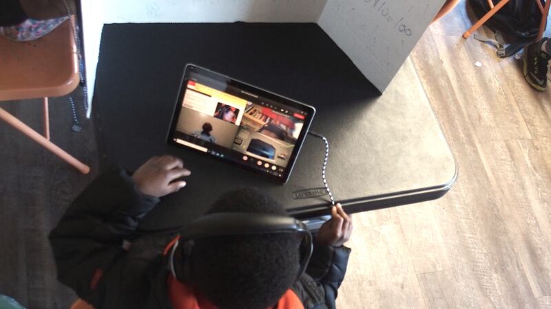 A young boy in a black and orange hoodie conducts virtual learning on his computer, sitting at a black partitioned desk.
