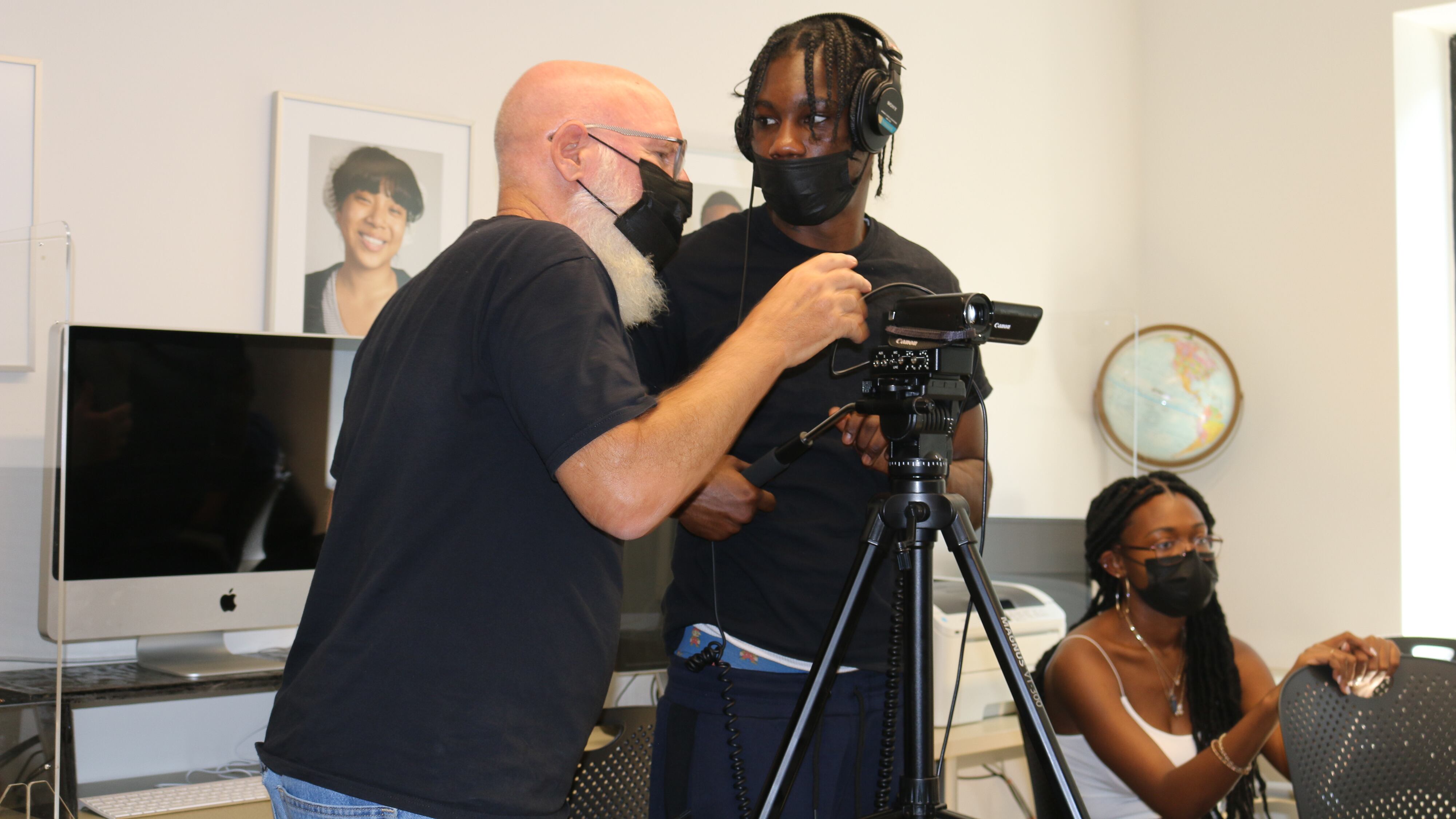 A man in a mask leans over a video camera while a student stands beside him.