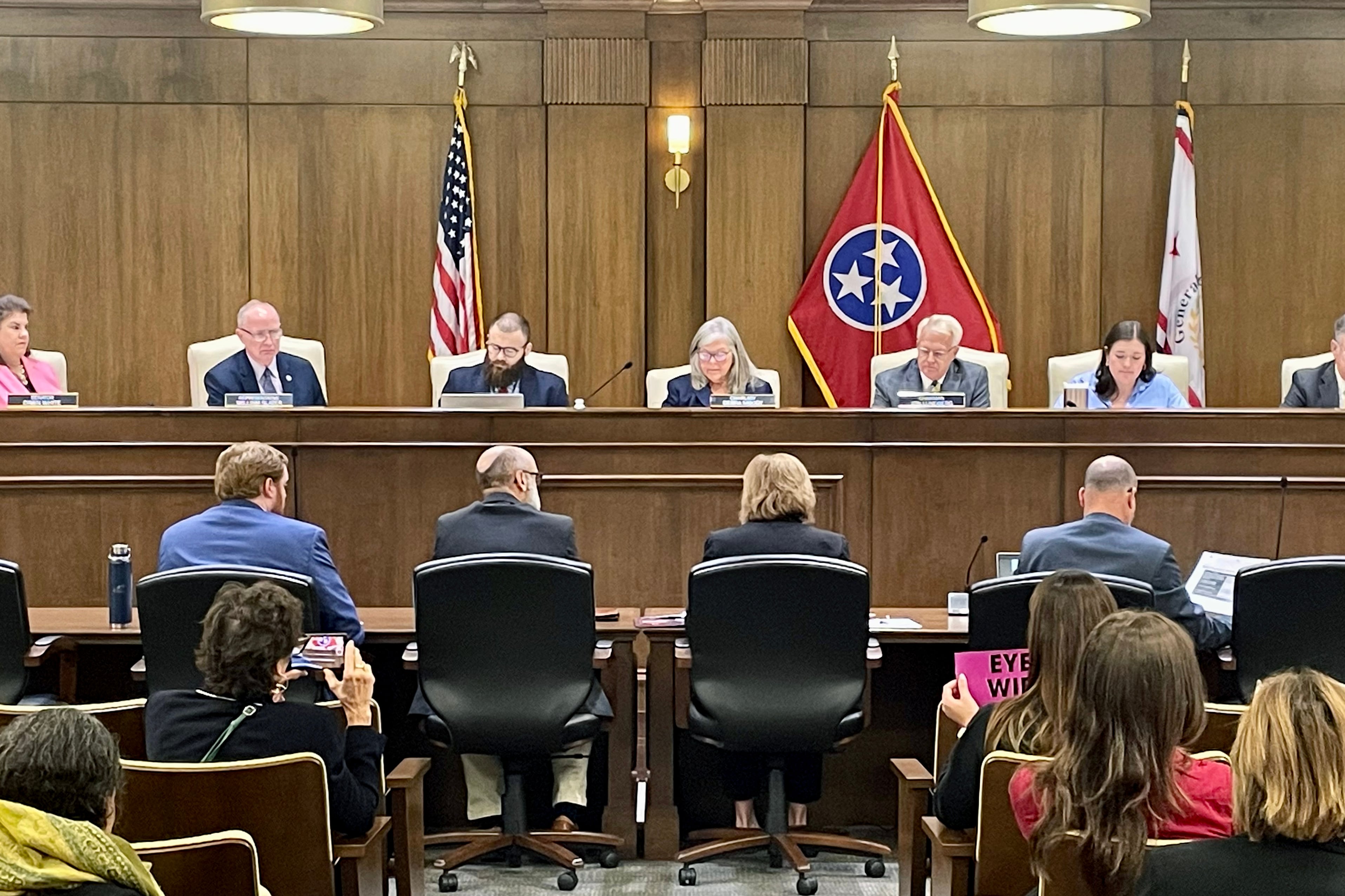 Seven people sit at a wooden court bench with flags in the background and people sitting in chairs in the foreground.