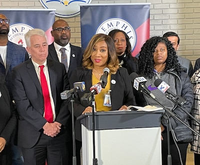 A woman with a dark suit jacket stands at a microphone surrounded by other people.