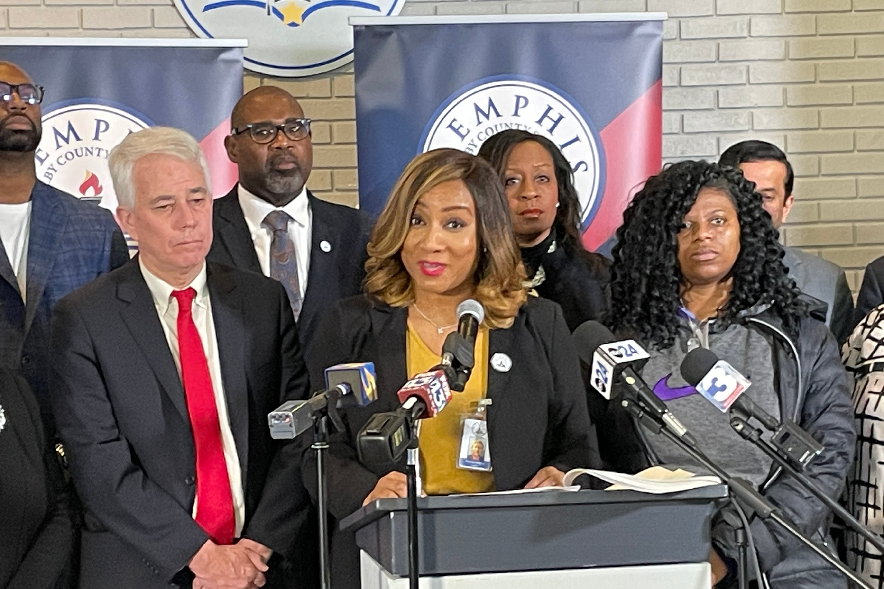 A woman with a dark suit jacket stands at a microphone surrounded by other people.
