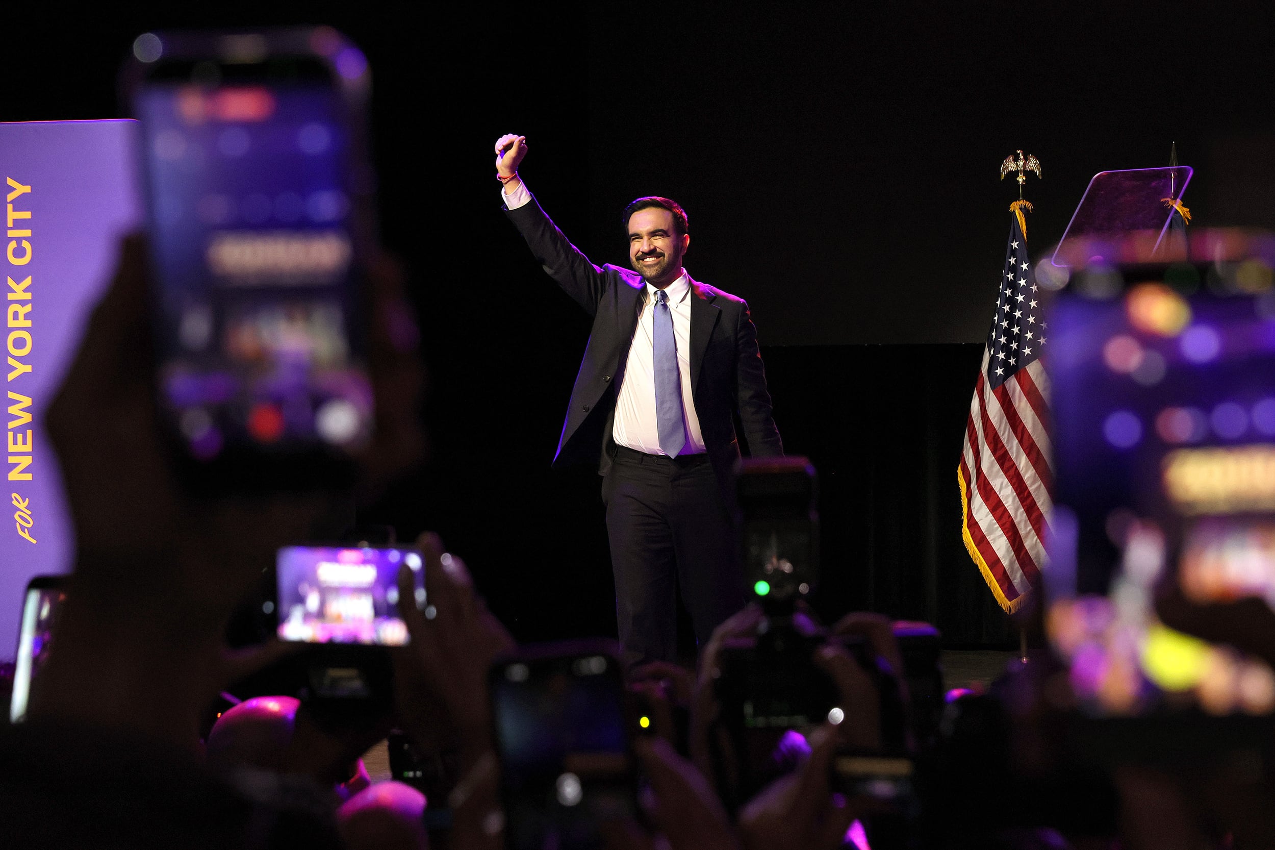 A photograph of a man in a suit with his fist in the air celebrating while walking onto a dark stage.